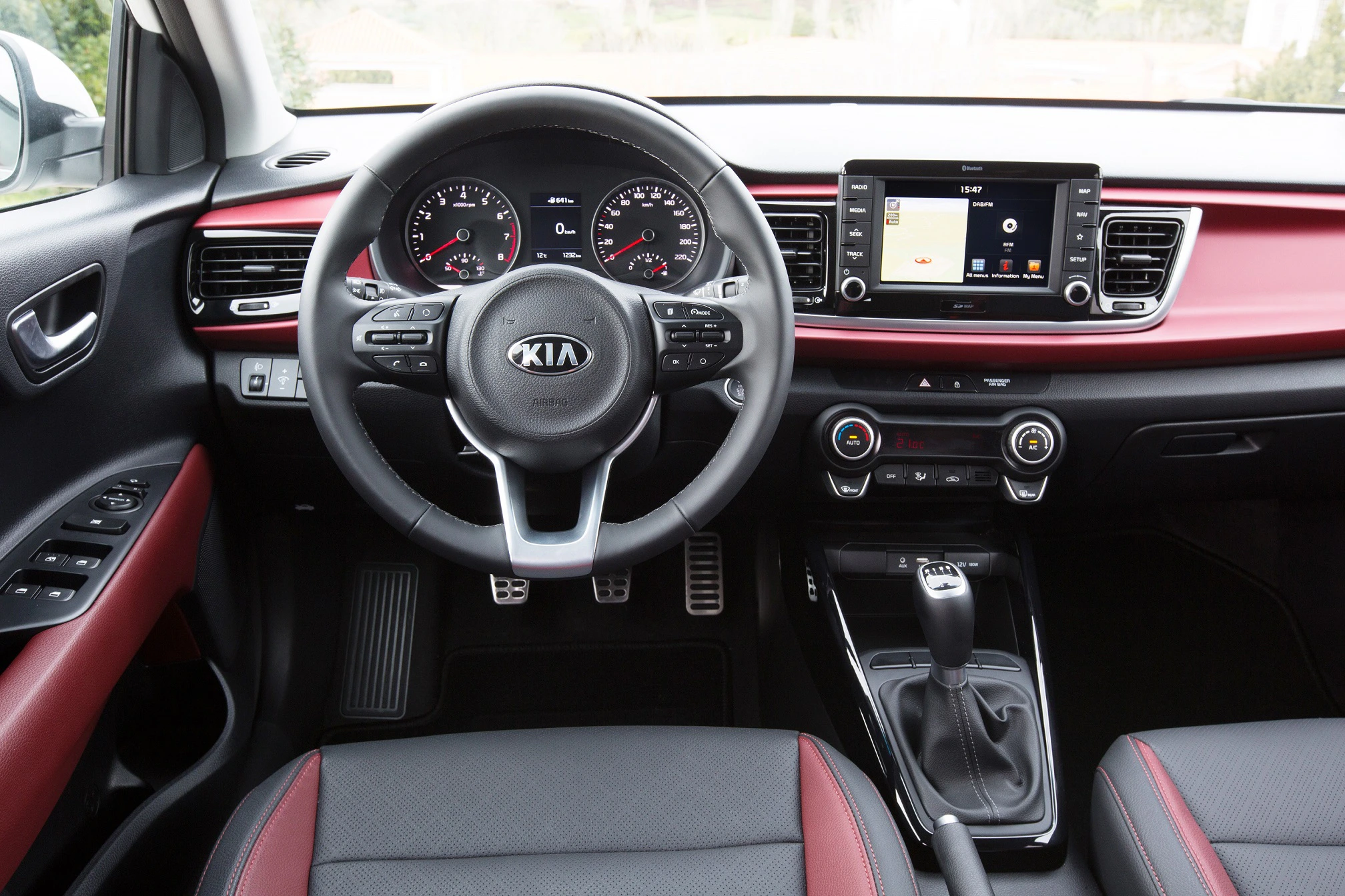 Innenraum eines Autos mit Lenkrad, Schalthebel, Tachometer und Infotainment-Bildschirm. Schwarze und rote Details an Armaturenbrett und Sitzen.