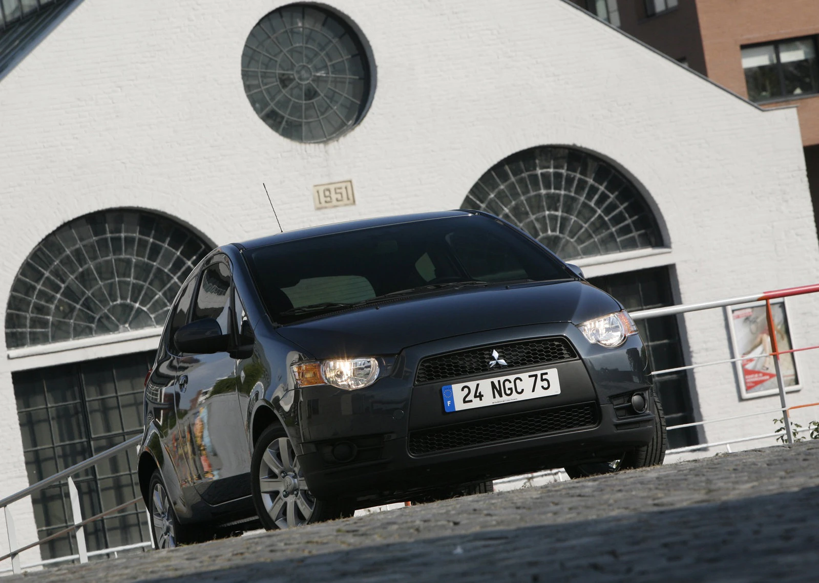 Schwarzer Mitsubishi Colt vor einem historischen Gebäude mit großen runden Fenstern und der Jahreszahl 1951 über dem Eingang.