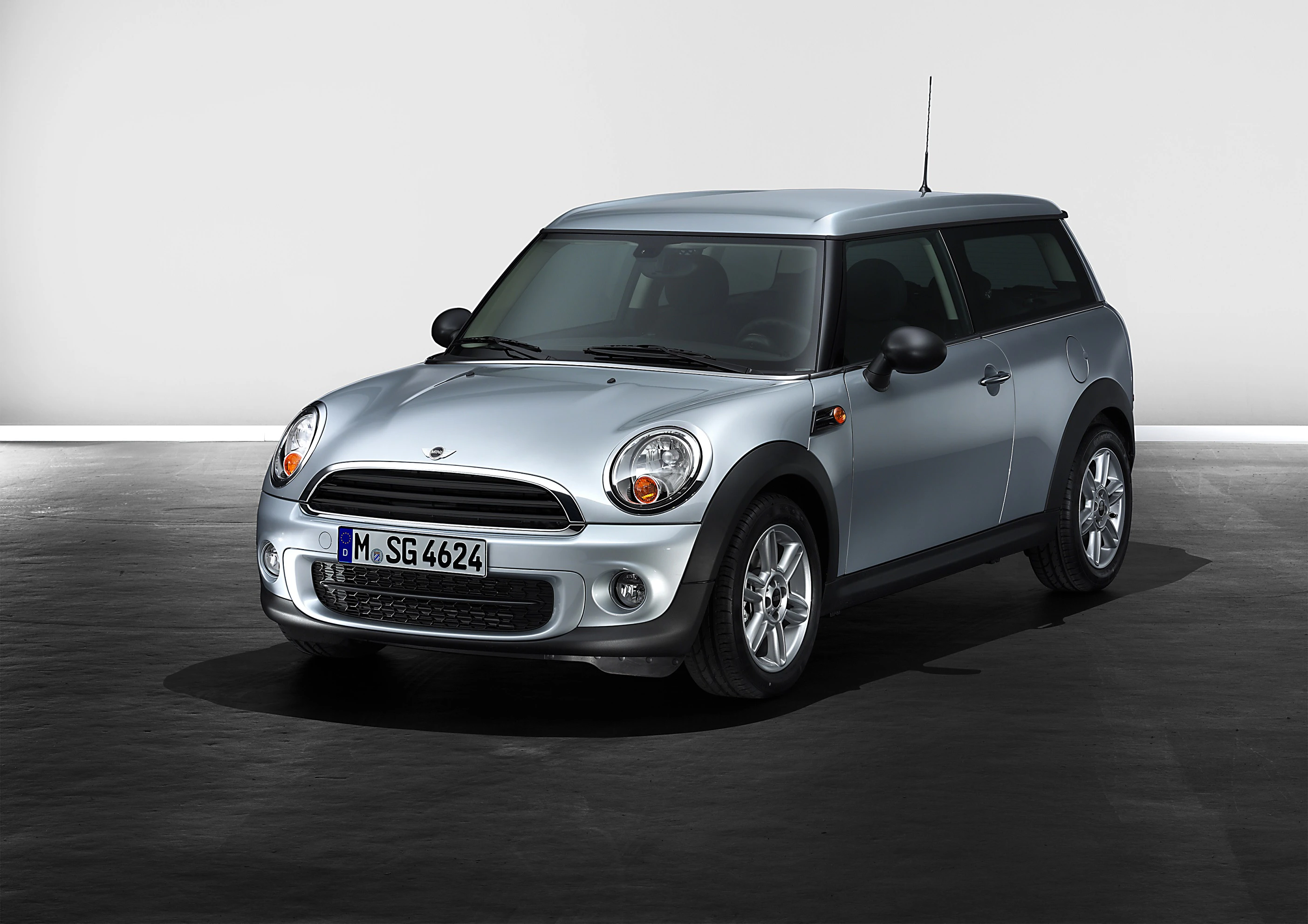 Silberner Mini Cooper Clubman steht auf dunklem Asphalt vor einer grauen Wand. Auto von vorne links sichtbar.