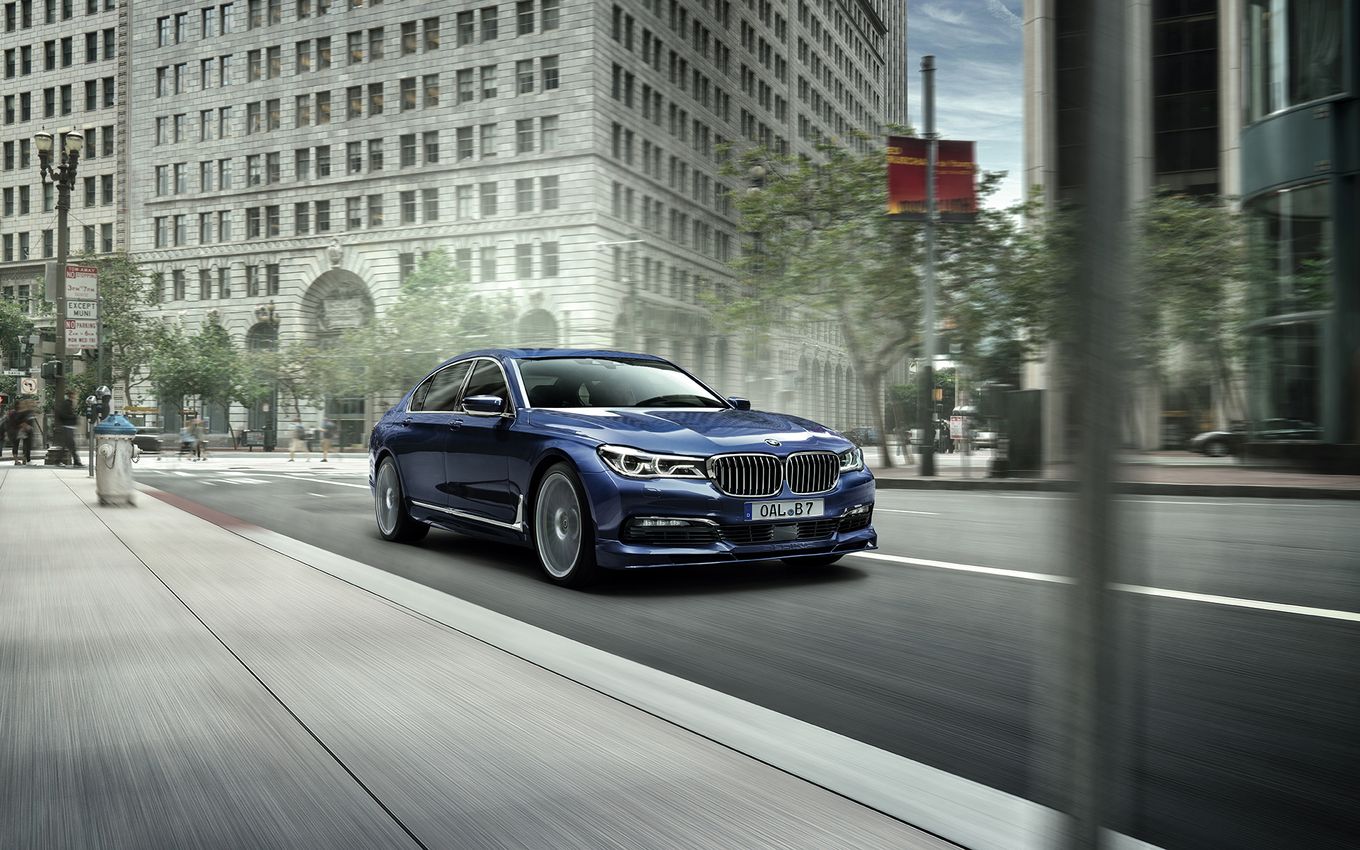 Blaues Auto fährt auf einer Stadtstraße, umgeben von hohen Gebäuden und Bäumen bei klarem Himmel.