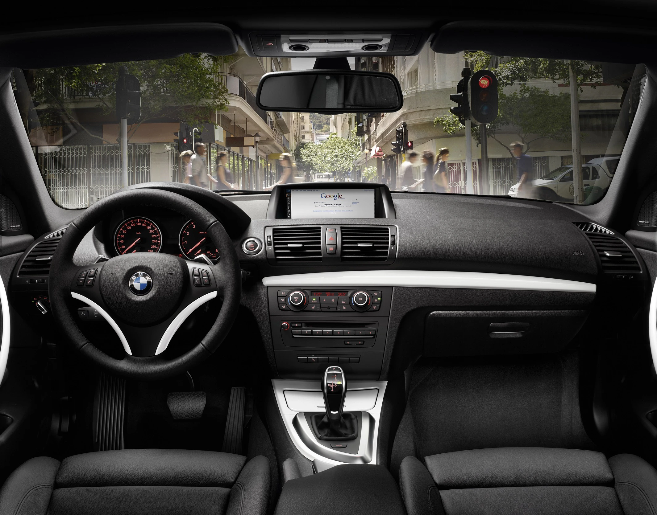 Innenraum eines BMW mit Lenkrad, Armaturenbrett, Gangschaltung und Navigationssystem. Blick auf städtische Straße durch die Frontscheibe.