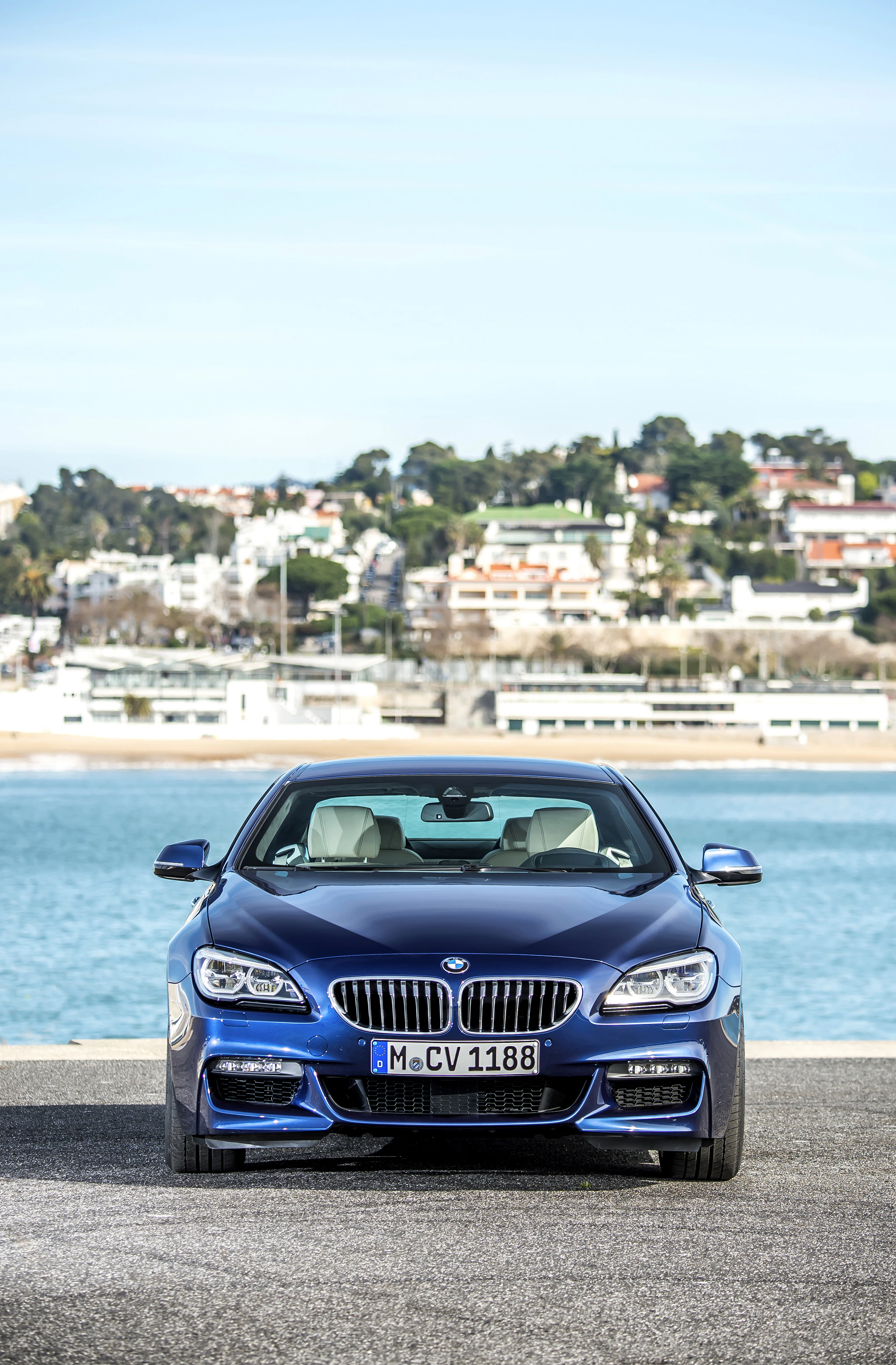 Blauer BMW steht auf einer Uferstraße, im Hintergrund Wasser und Stadt. Klarer Himmel und modern gestaltetes Fahrzeug.