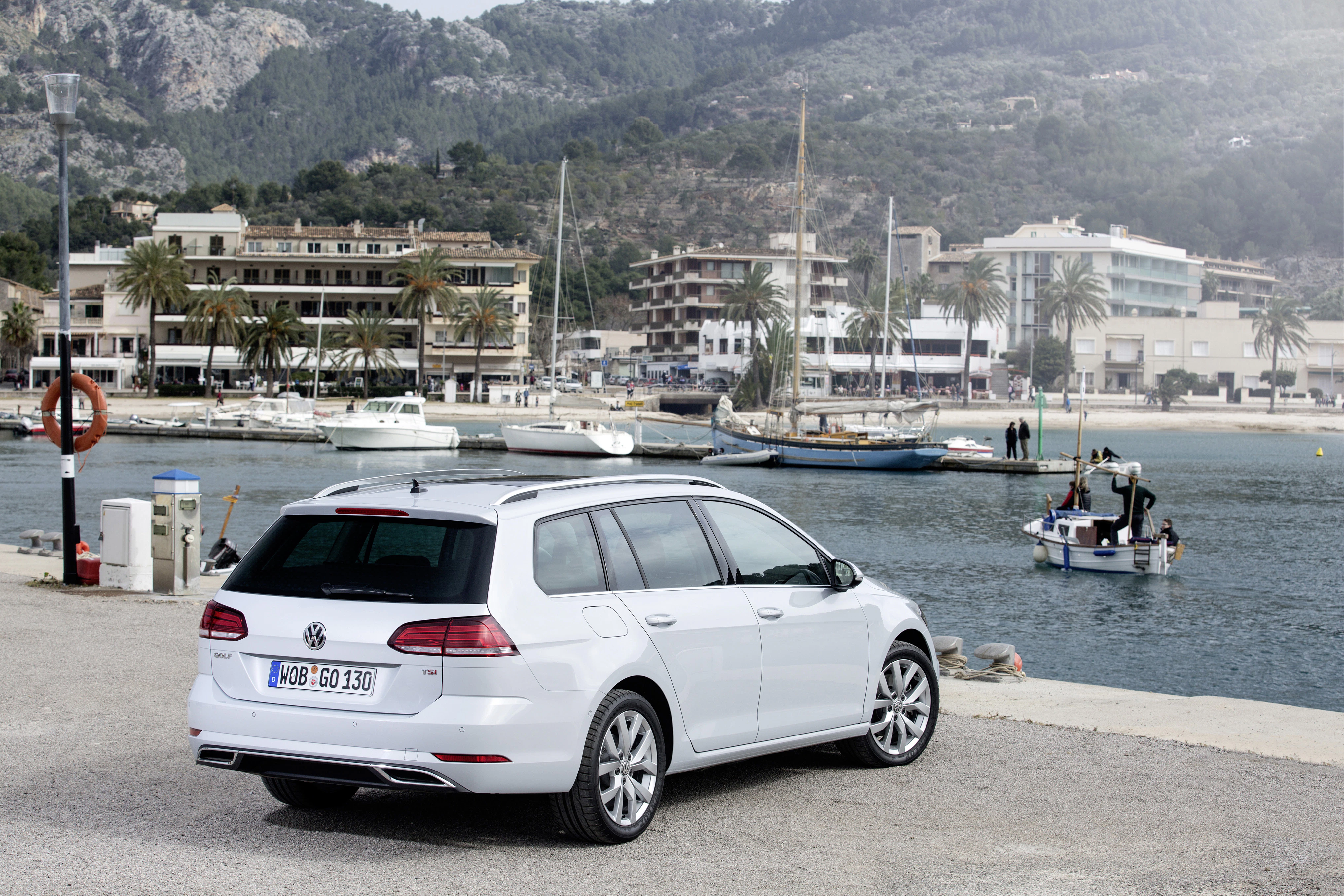 Ein weißer VW Golf Variant parkt vor einer malerischen Hafenlandschaft mit Booten und Bergen im Hintergrund.
