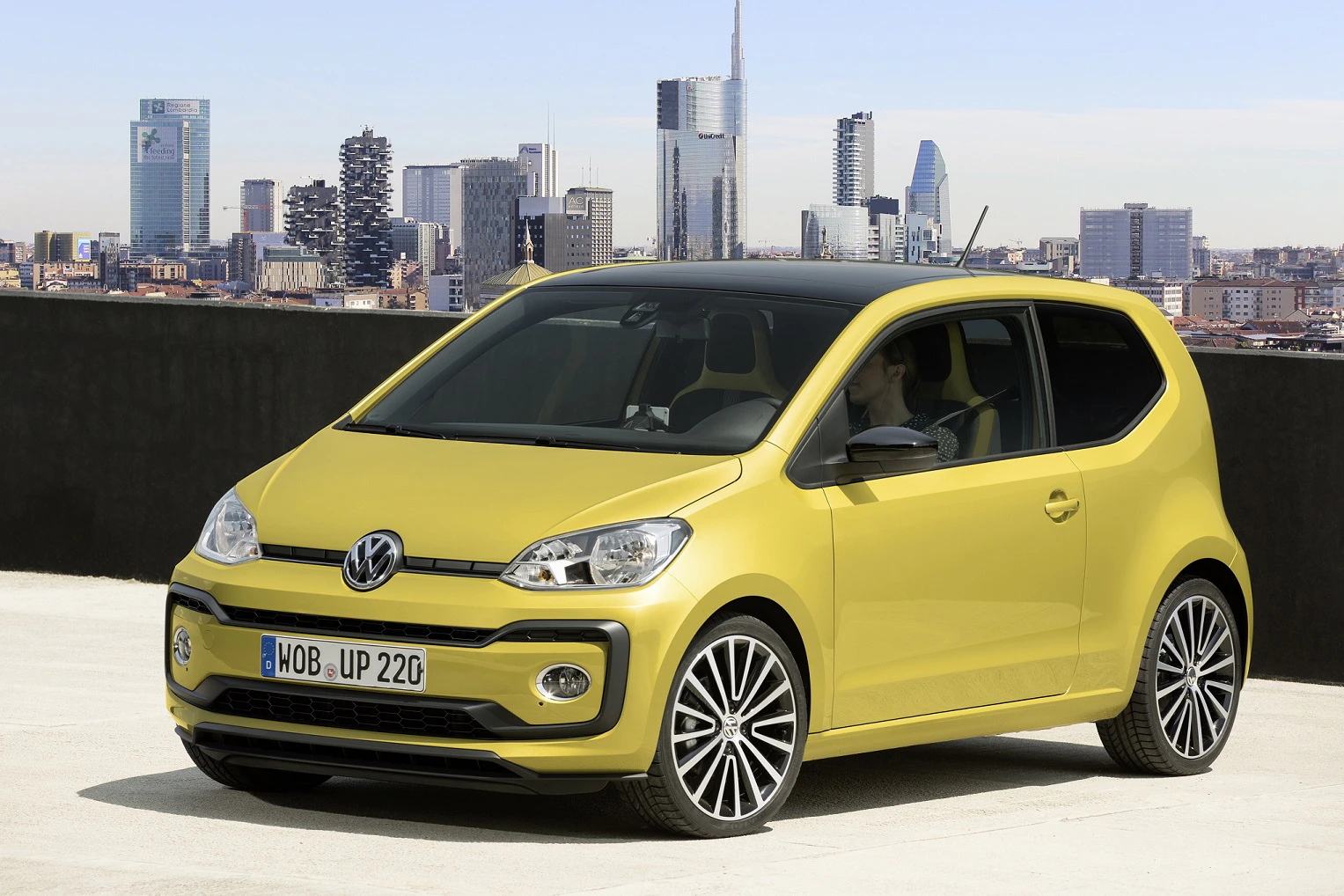 Gelbes Volkswagen-Auto auf einem Parkplatz mit Stadtpanorama im Hintergrund. Moderne Gebäude und klarer Himmel sind sichtbar.