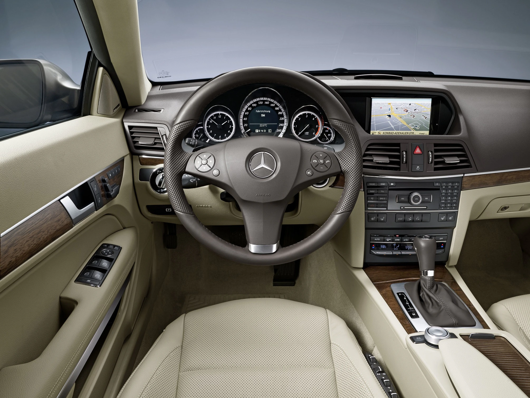 Innenraum eines Mercedes mit beigem Leder, Holzverkleidung, Lenkrad mit Logo, Armaturenbrett mit Anzeige, Touchscreen und Automatikschaltung.