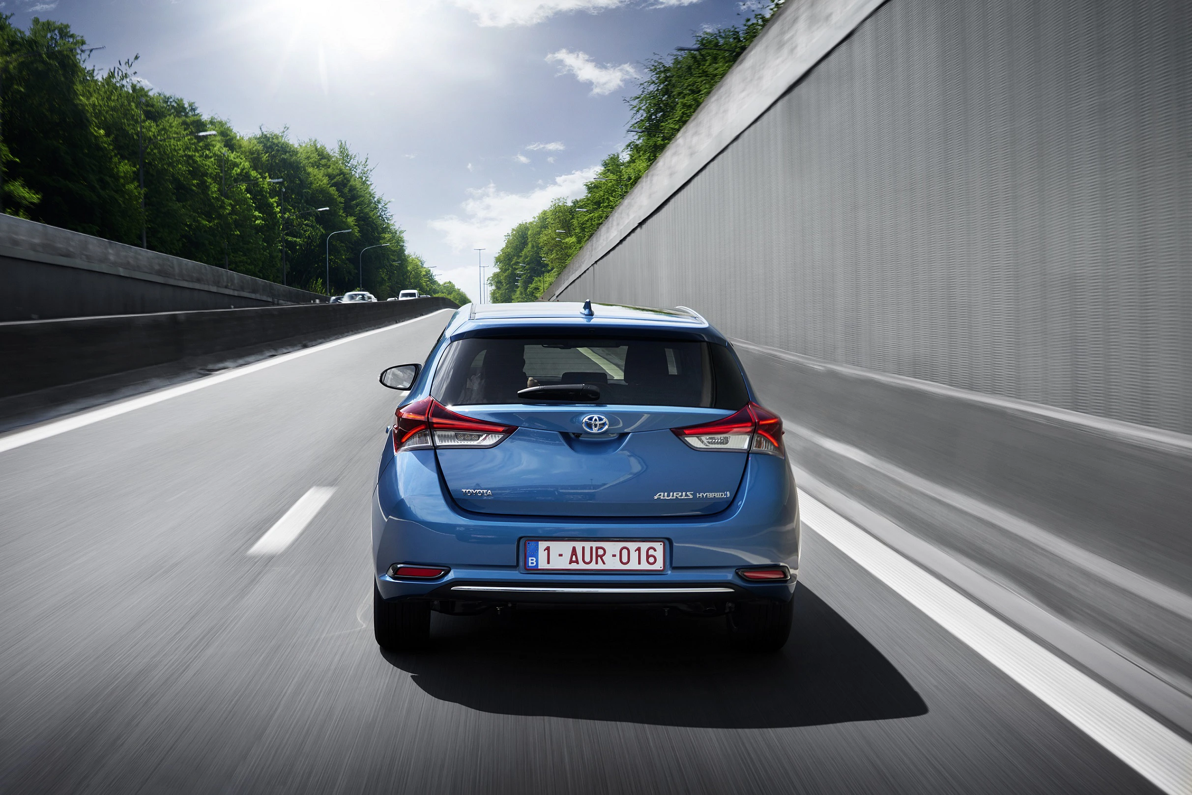 Blauer Toyota Auris fährt auf einer leeren Autobahn, umgeben von Bäumen und einer hohen Mauer auf der rechten Seite.