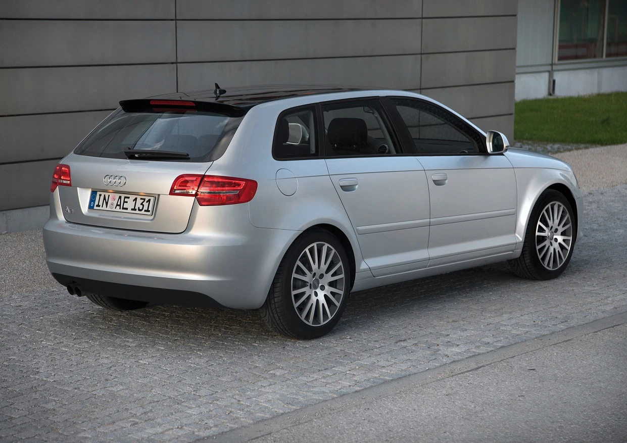Silberner Audi A3 Sportback, geparkt auf Kopfsteinpflaster vor einem modernen Gebäude, Ansicht von hinten links.