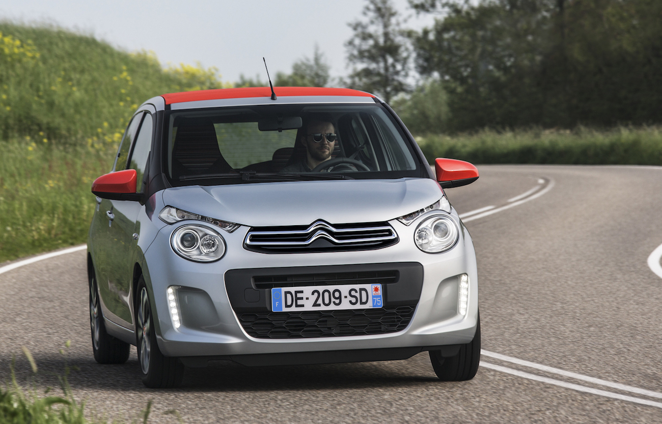 Silberner Citroën mit rotem Dach fährt auf gewundener Landstraße; Fahrer mit Sonnenbrille am Steuer.