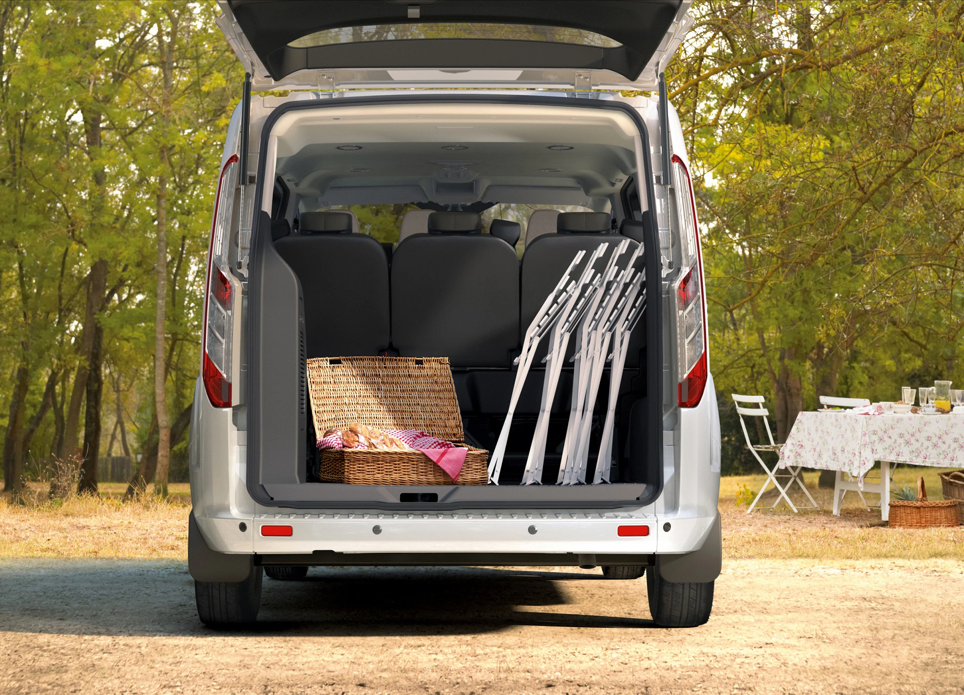 Geöffneter Kofferraum eines Vans mit Picknickkorb und gefalteten Stühlen. Im Hintergrund ein gedeckter Tisch im Grünen.