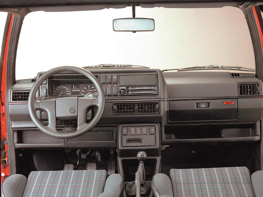 Innenraum eines Oldtimer-Autos mit schwarzem Armaturenbrett, Lenkrad, Schalthebel und karierten Sitzen. Vintage-Design-Elemente sichtbar.