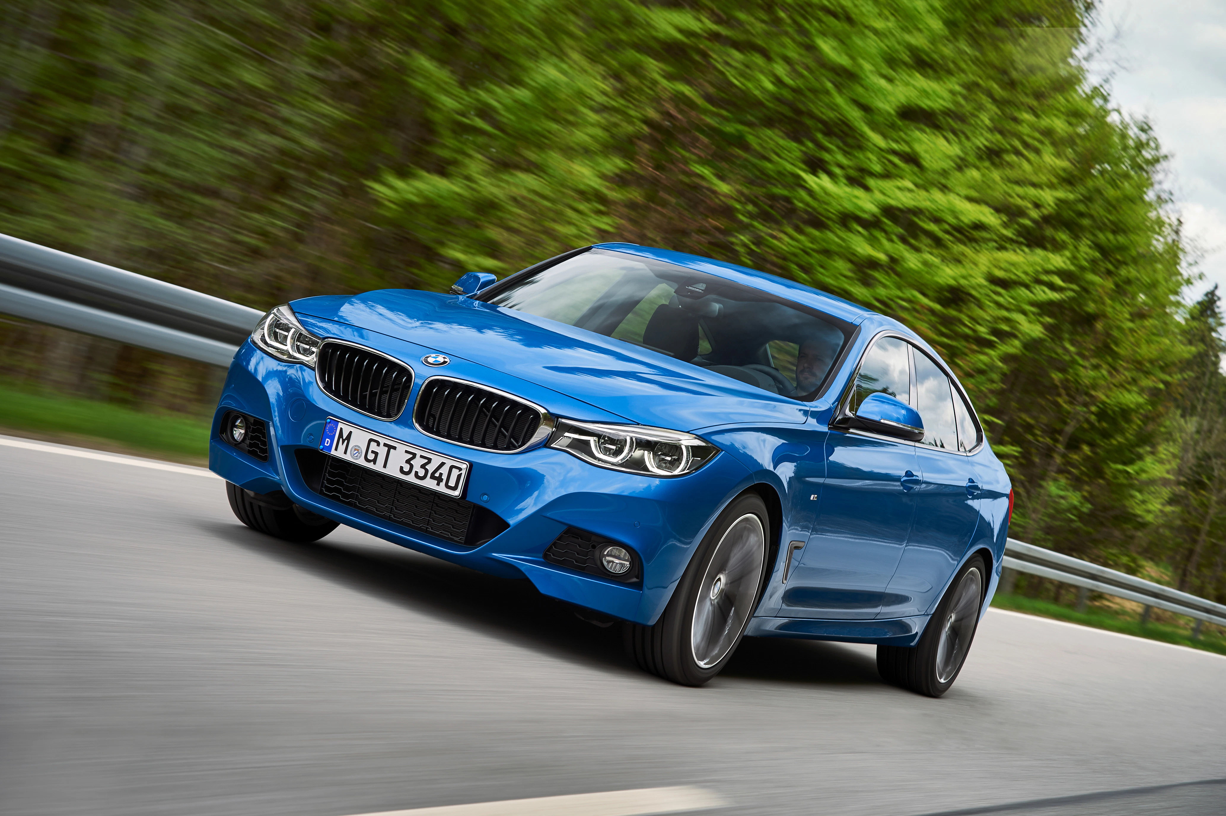Blauer BMW auf einer Landstraße, umgeben von grünen Bäumen, fährt schnell. Dynamische Frontansicht mit sichtbarem Nummernschild.