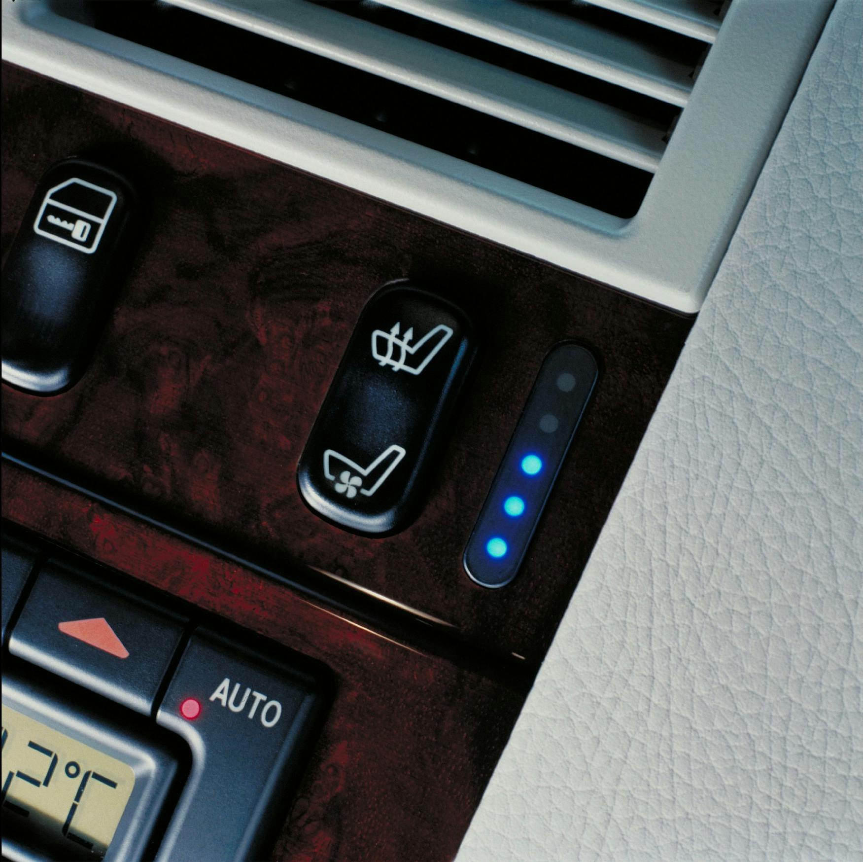 Nahaufnahme eines Autositz-Heizungsreglers mit blauen LED-Anzeigen und Holzdekorverkleidung. Lüftungsschlitze sind oben sichtbar.