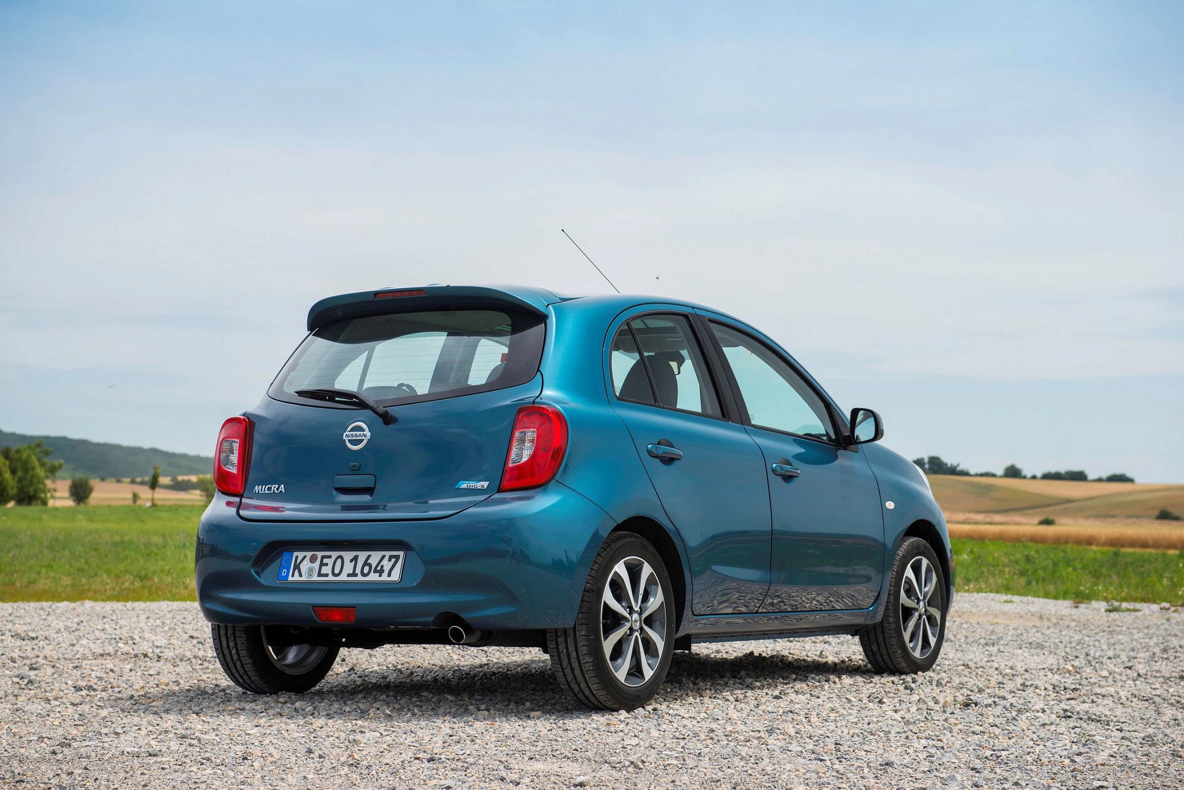 Blauer Kleinwagen steht auf einem Schotterplatz, im Hintergrund Wiesen und blauer Himmel.