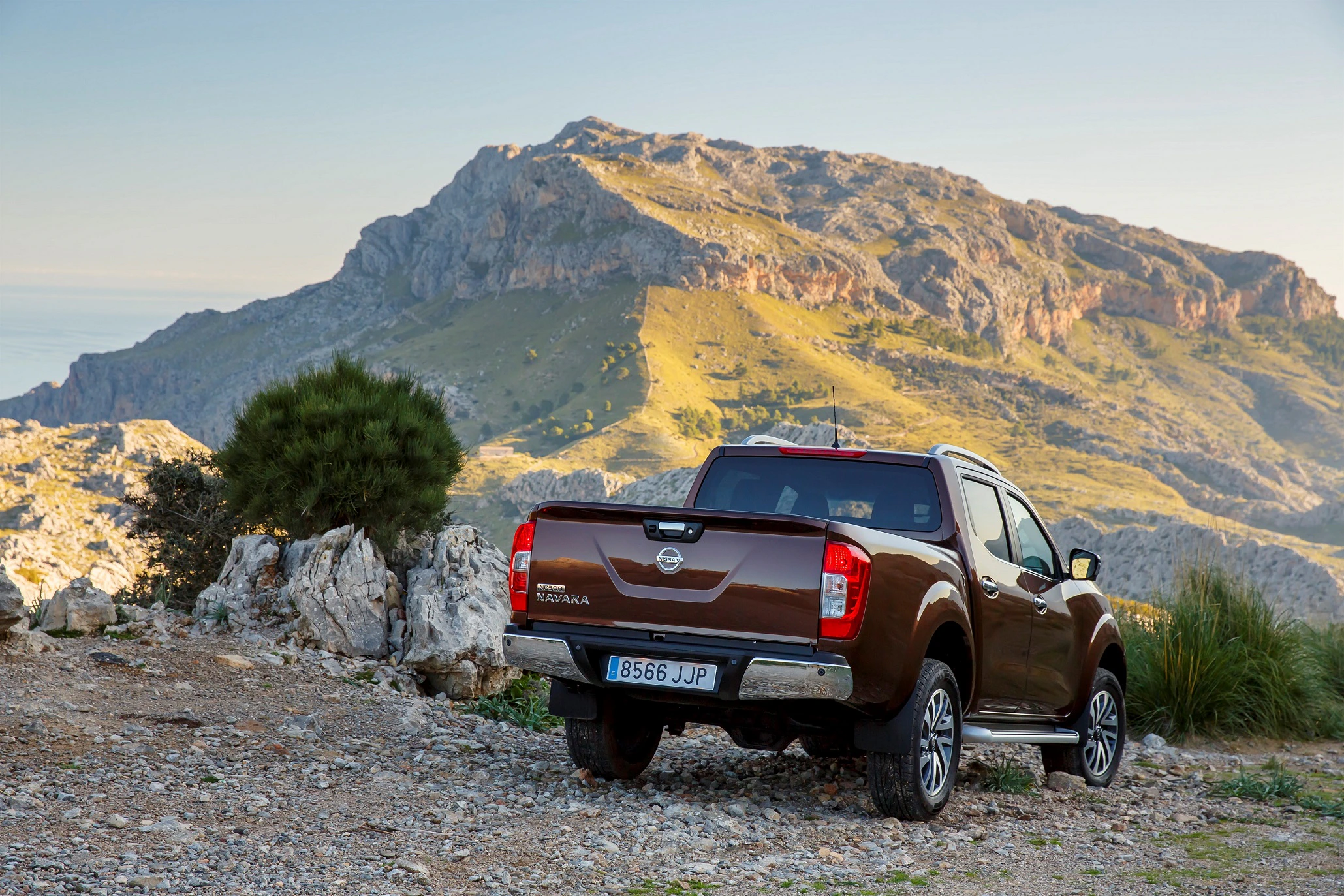 Brauner Pickup auf steinigem Weg vor einer hügeligen Landschaft mit blauem Himmel im Hintergrund. Nummernschild sichtbar.