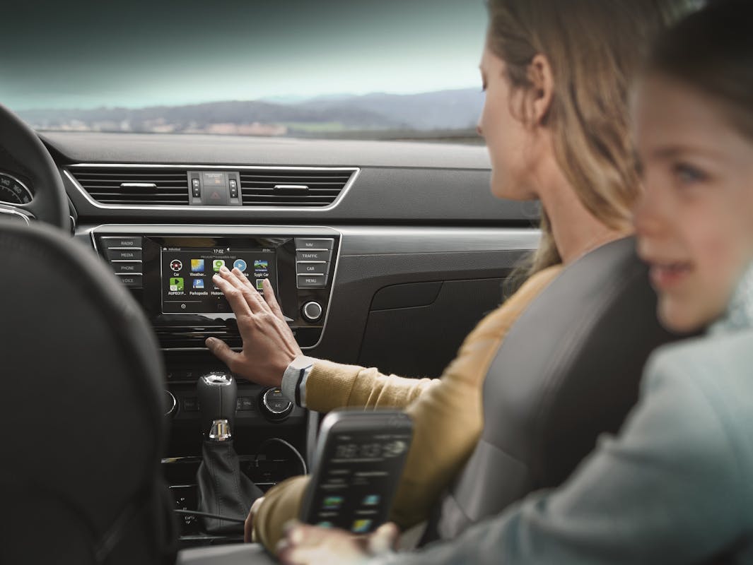 Eine Frau bedient den Touchscreen im Auto, während sie neben einem Kind auf dem Rücksitz sitzt. Autoinnenraum im Hintergrund sichtbar.