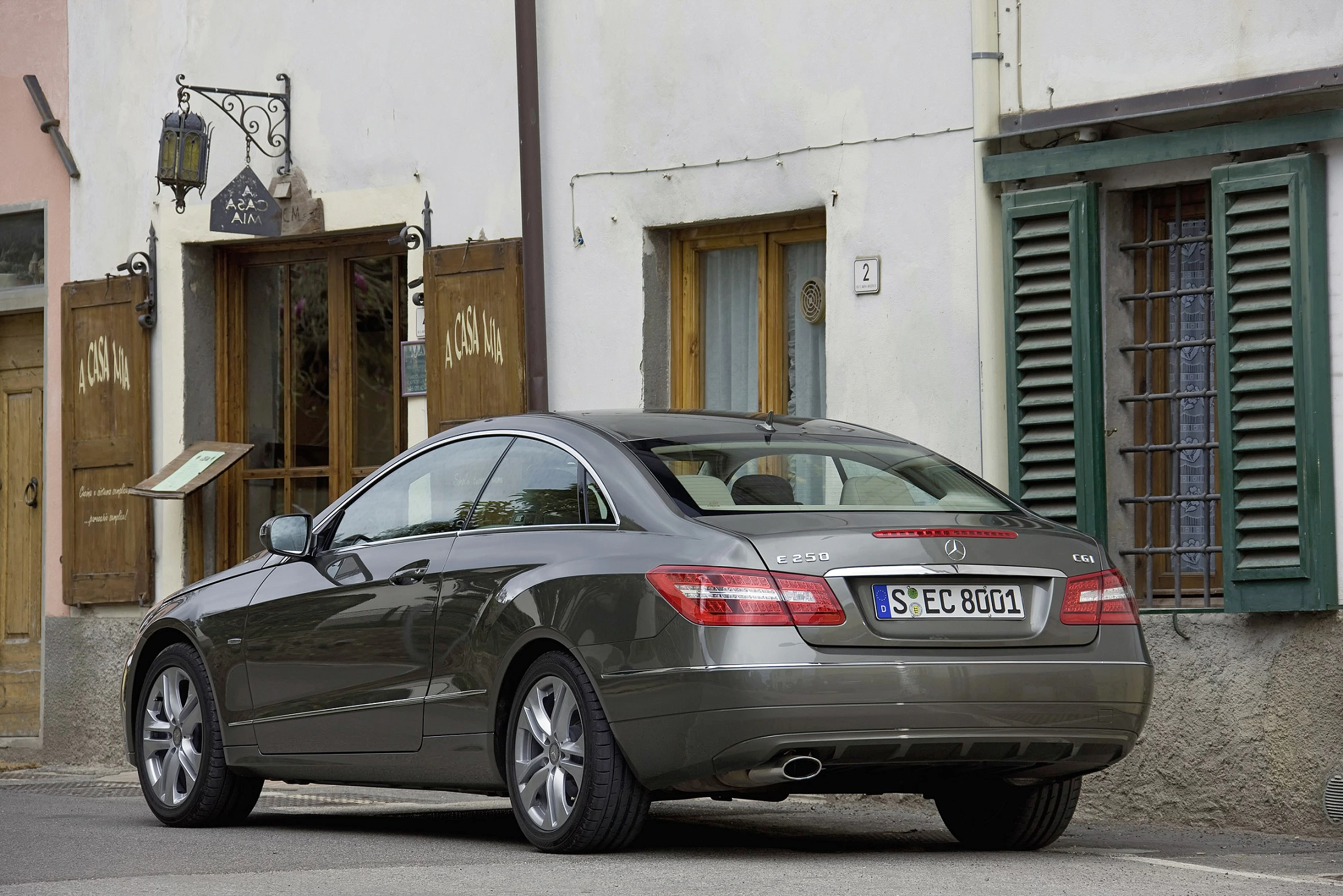 Graues Mercedes-Benz Coupé parkt vor einem Gebäude mit grünen Fensterläden und Holztüren in einer europäischen Straße.