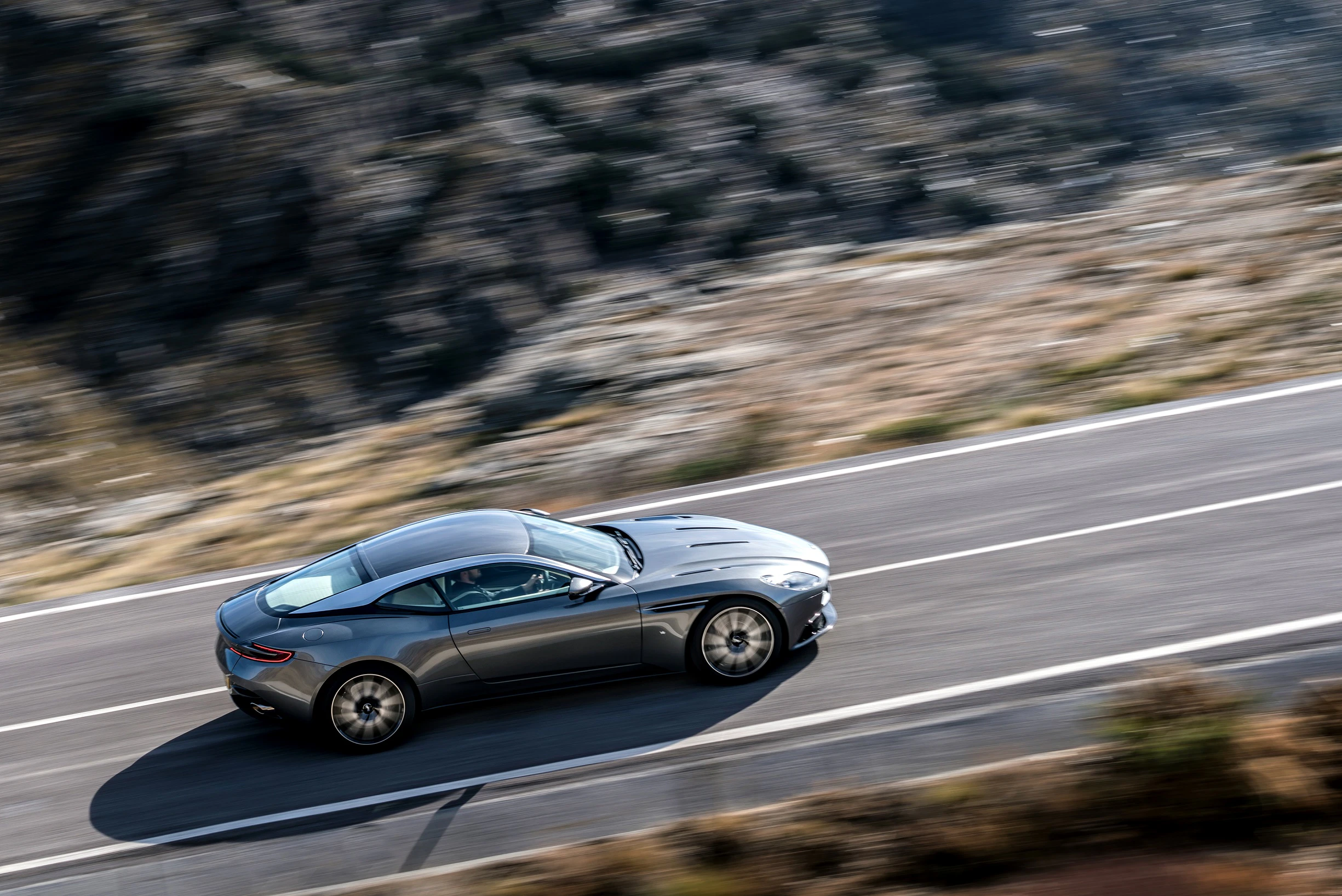 Graues Sportcoupé fährt auf kurvenreicher Bergstraße, umgeben von felsiger Landschaft und unscharfem Hintergrund.