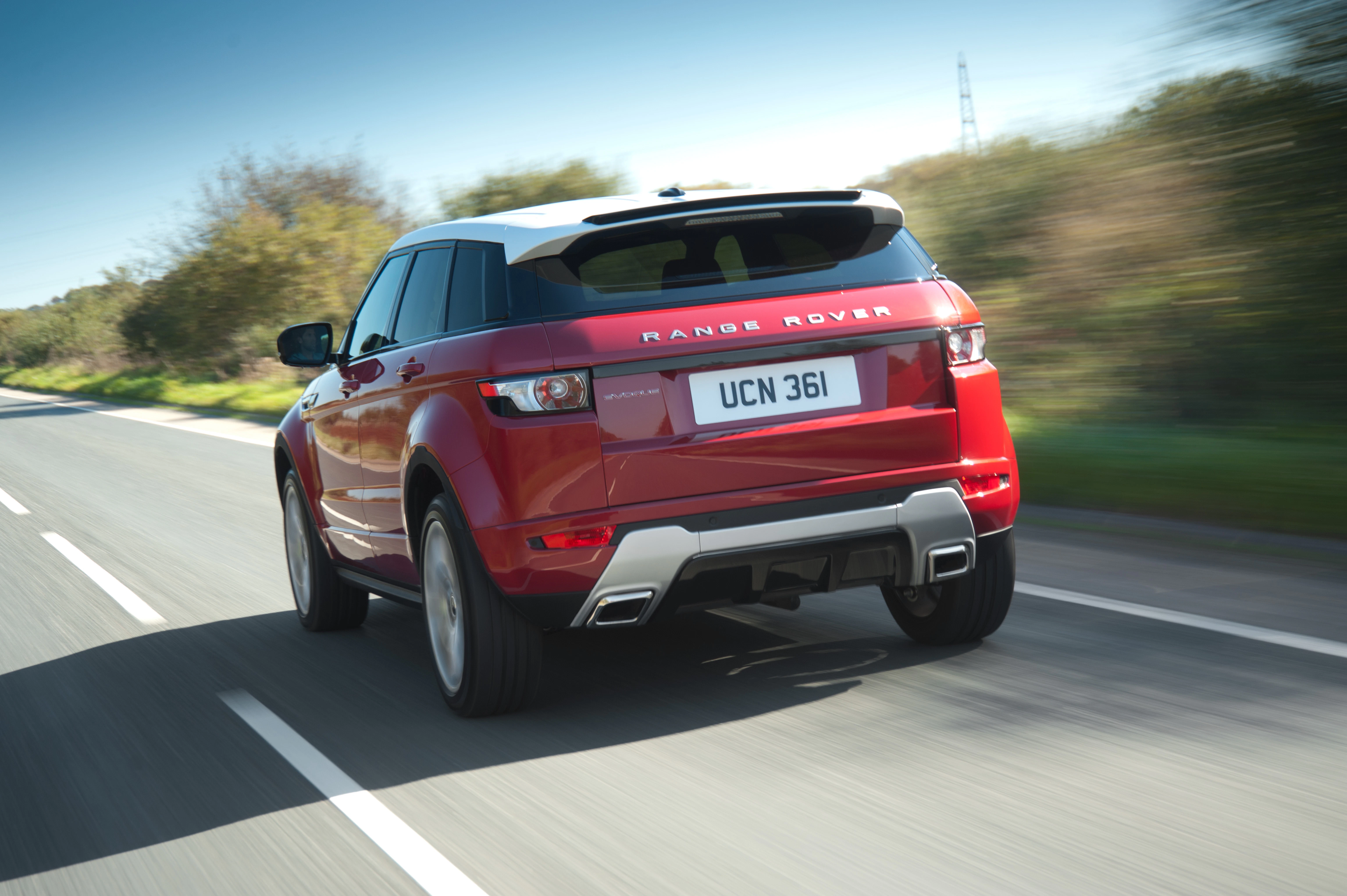 Roter Range Rover Evoque fährt auf einer Landstraße bei sonnigem Wetter, umgeben von Bäumen und blauem Himmel.