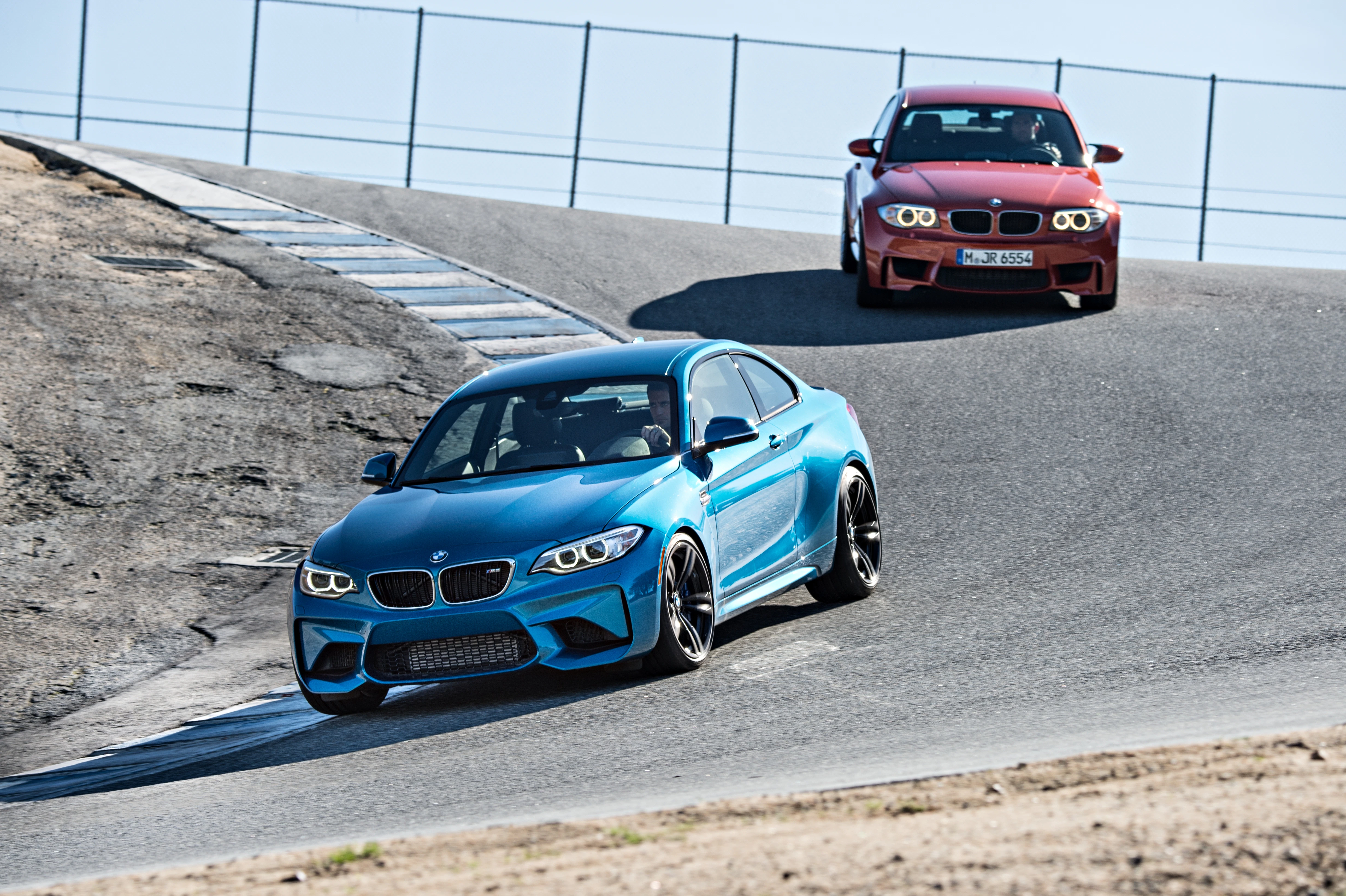 Zwei BMWs auf einer Rennstrecke, ein blauer vorne in einer Kurve, ein roter dahinter. Sonniger Tag, Reifenprüfspuren sichtbar.