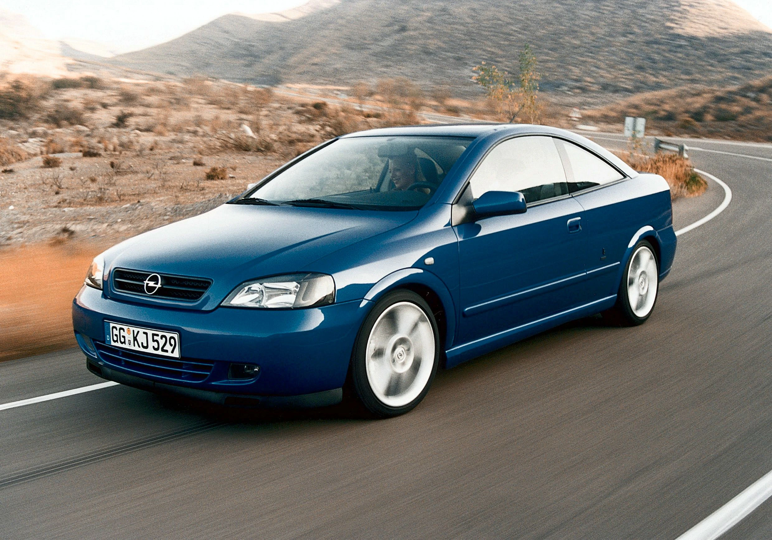 Blauer Opel-Coupé fährt auf kurviger Straße in karger Landschaft. Geschwindigkeit und Eleganz im Fokus.