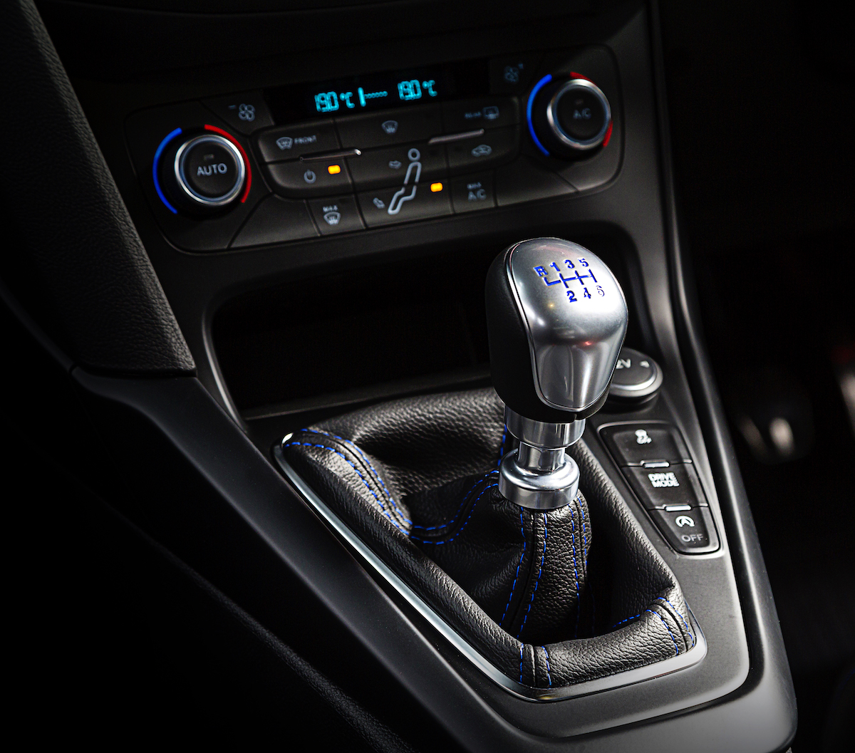 Nahaufnahme eines Autoschaltknüppels mit Ledermanschette und blauen Nähten, umgeben von Bedienelementen für Heizung und Klima.