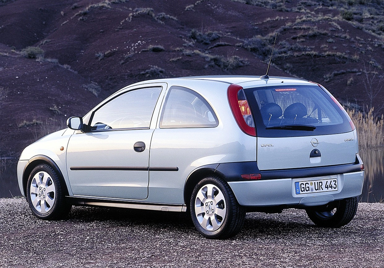 Silberner dreitüriger Kleinwagen, Opel Corsa, geparkt vor einem hügeligen Landschaftshintergrund. Rückansicht mit sichtbarem Nummernschild.