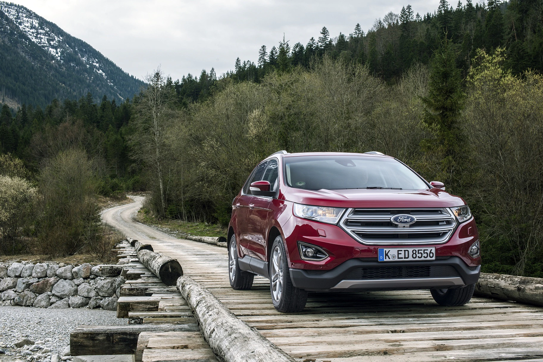 Roter SUV auf einer Holzbrücke in einer ländlichen Berglandschaft mit bewaldeten Hügeln und schneebedeckten Gipfeln im Hintergrund.
