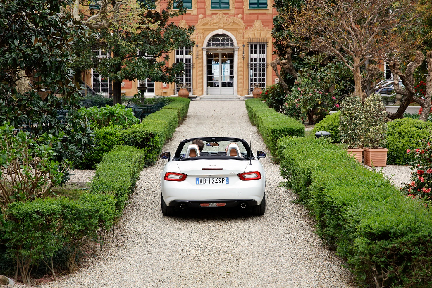 Weißes Cabrio auf einem Kiesweg, der zu einer eleganten Villa mit üppigen Büschen und Bäumen führt.