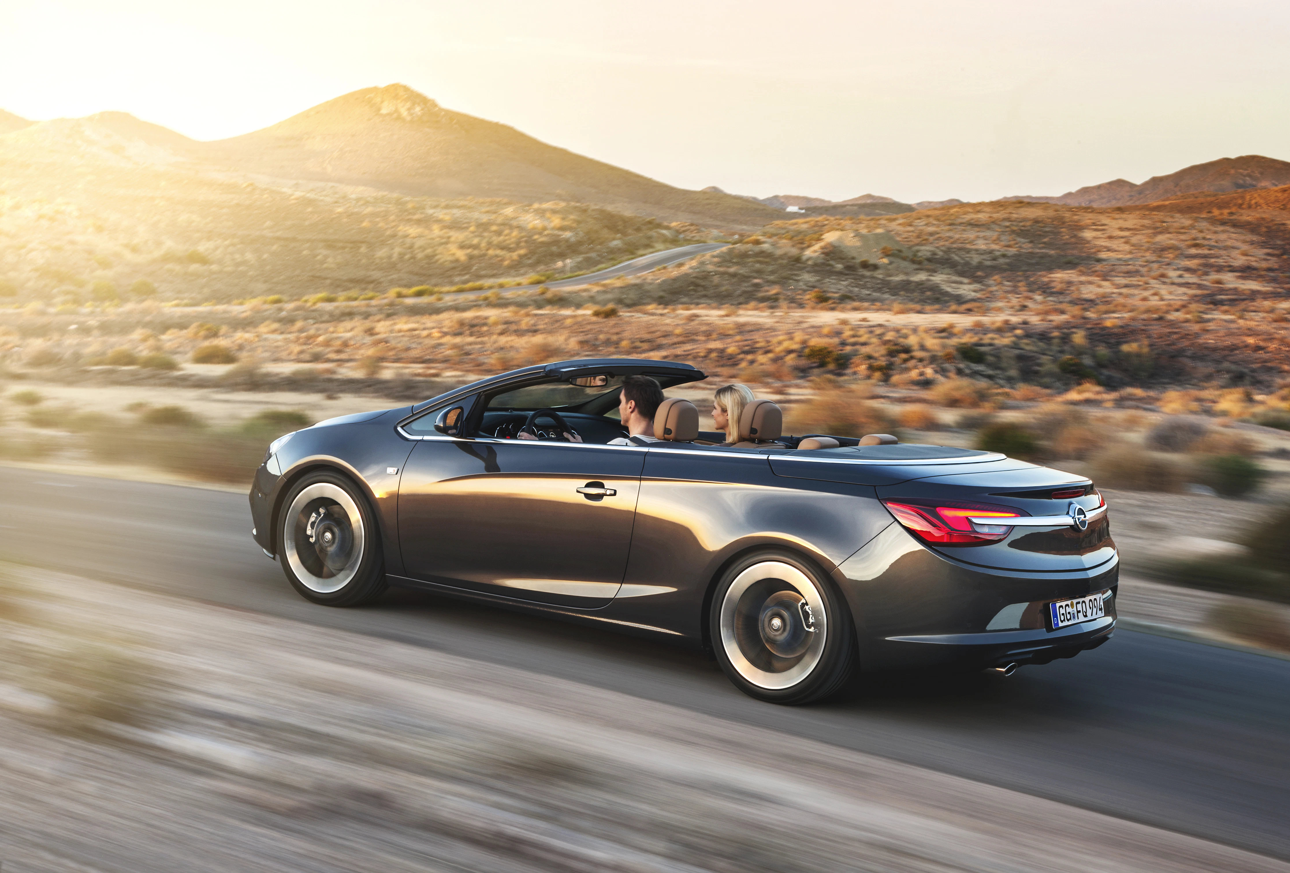 Ein Cabriolet fährt auf einer Straße durch eine bergige, sonnige Landschaft. Der Himmel ist klar und das Auto hat ein offenes Verdeck.