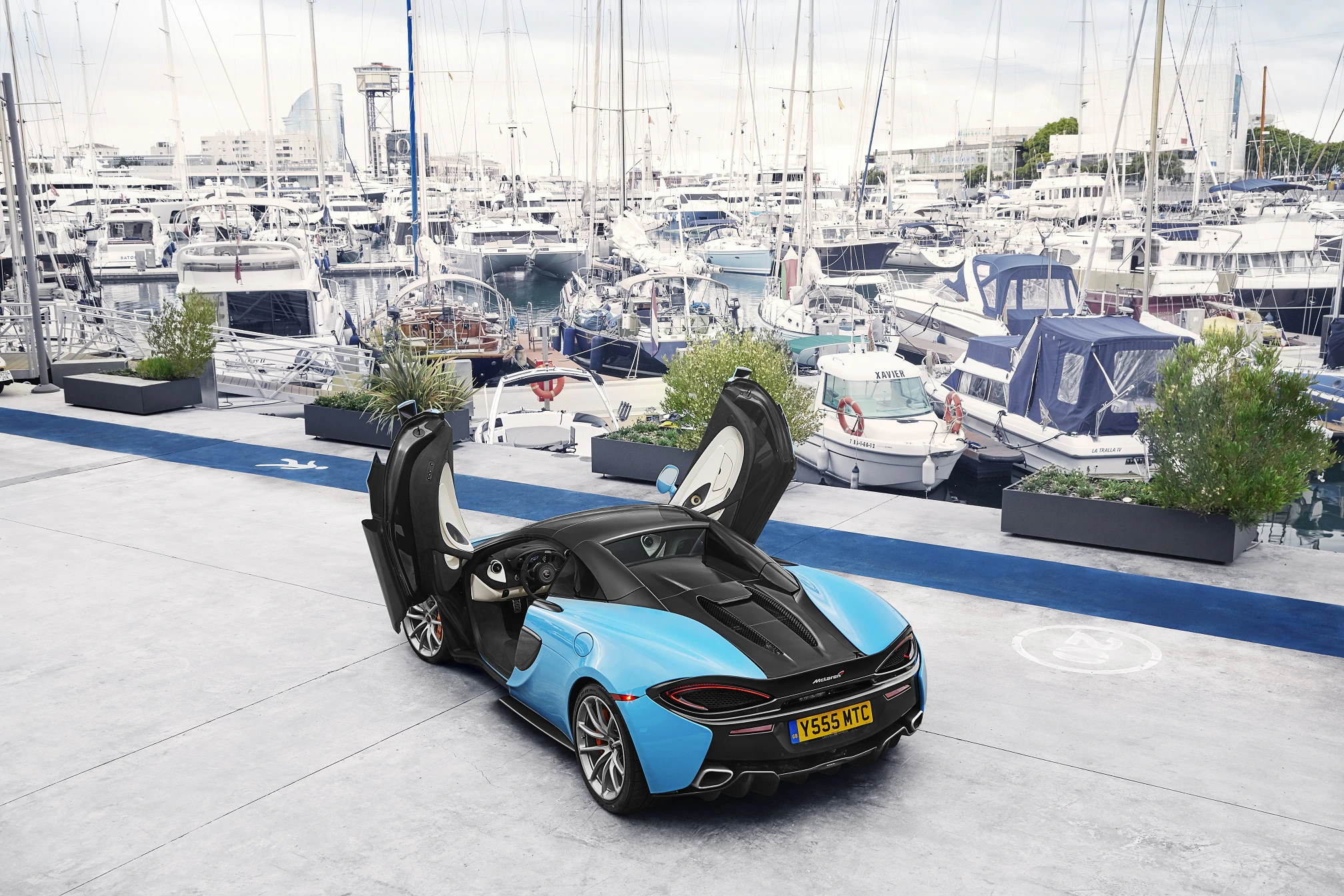 Blauer Sportwagen mit offenen Flügeltüren steht auf einem Parkplatz vor einem Yachthafen voller Boote.