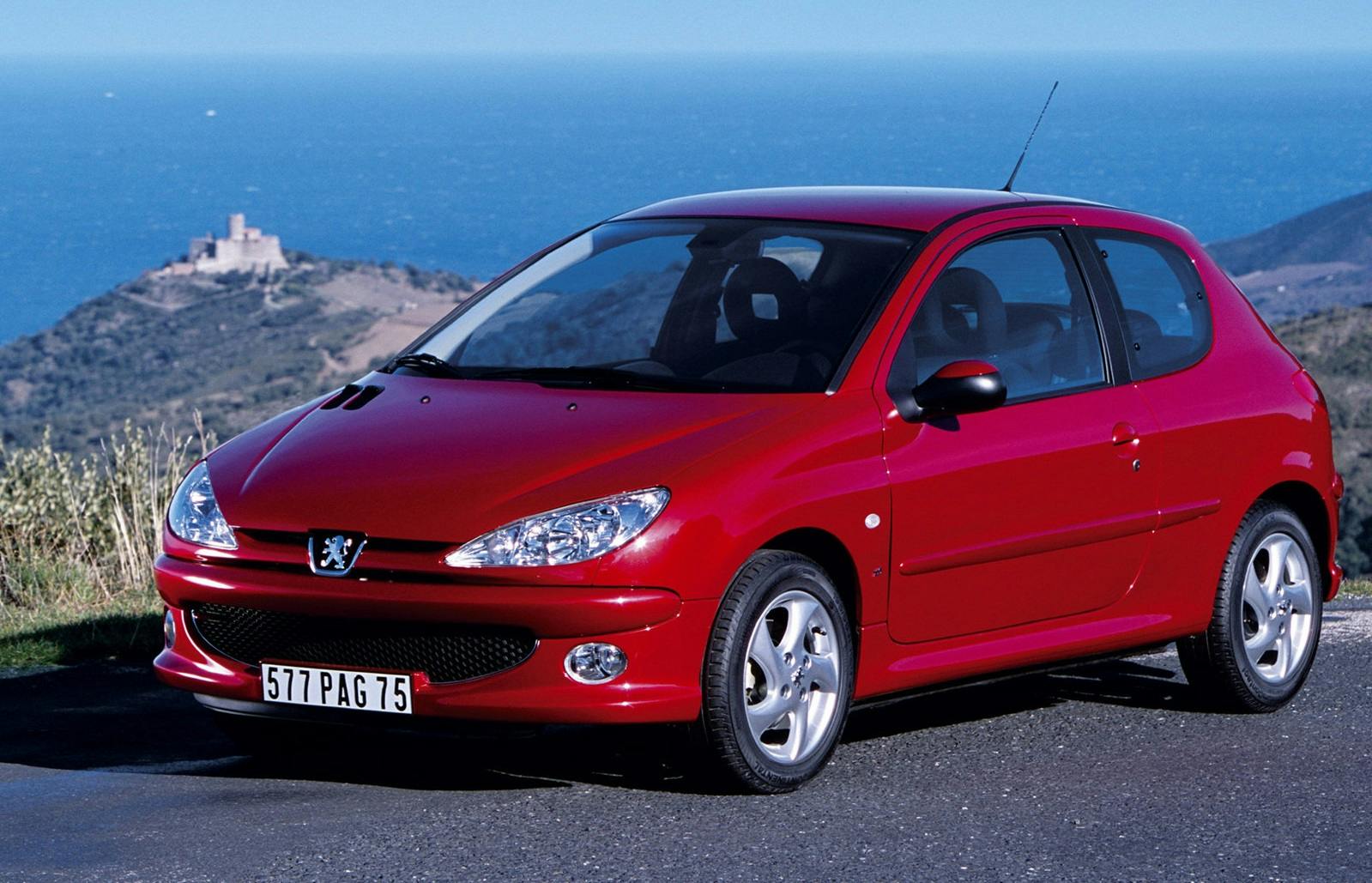 Roter Peugeot 206 auf einer Straße am Meer, mit einer Burg im Hintergrund unter klarem blauen Himmel.