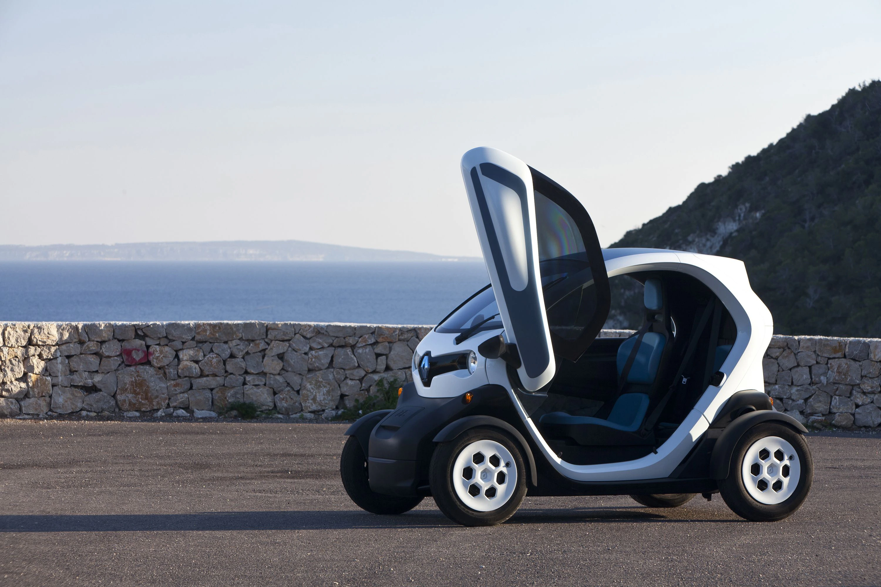 Kleines weißes Elektroauto mit offenen Flügeltüren, geparkt vor einer Steinmauer; im Hintergrund das Meer und bewaldete Hügel.