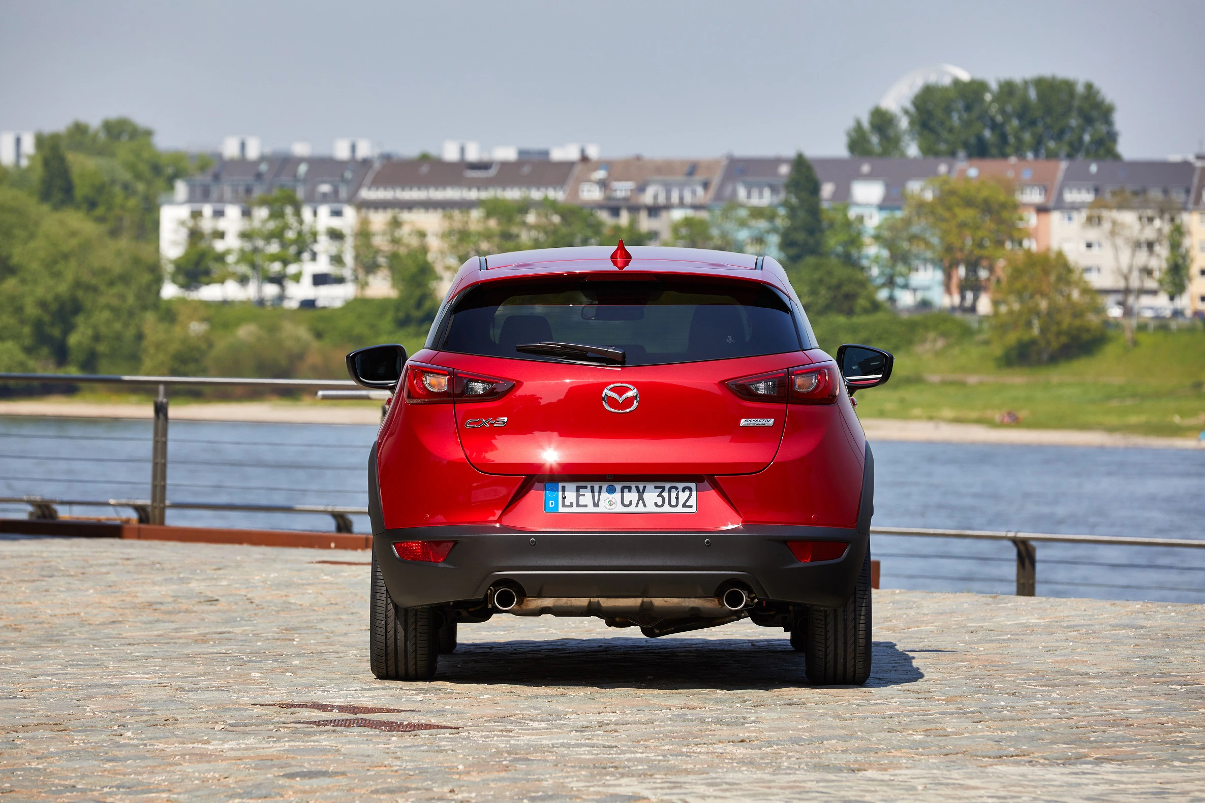 Roter SUV Mazda CX-5 von hinten, geparkt an einem Fluss mit Stadt im Hintergrund, sonniges Wetter.