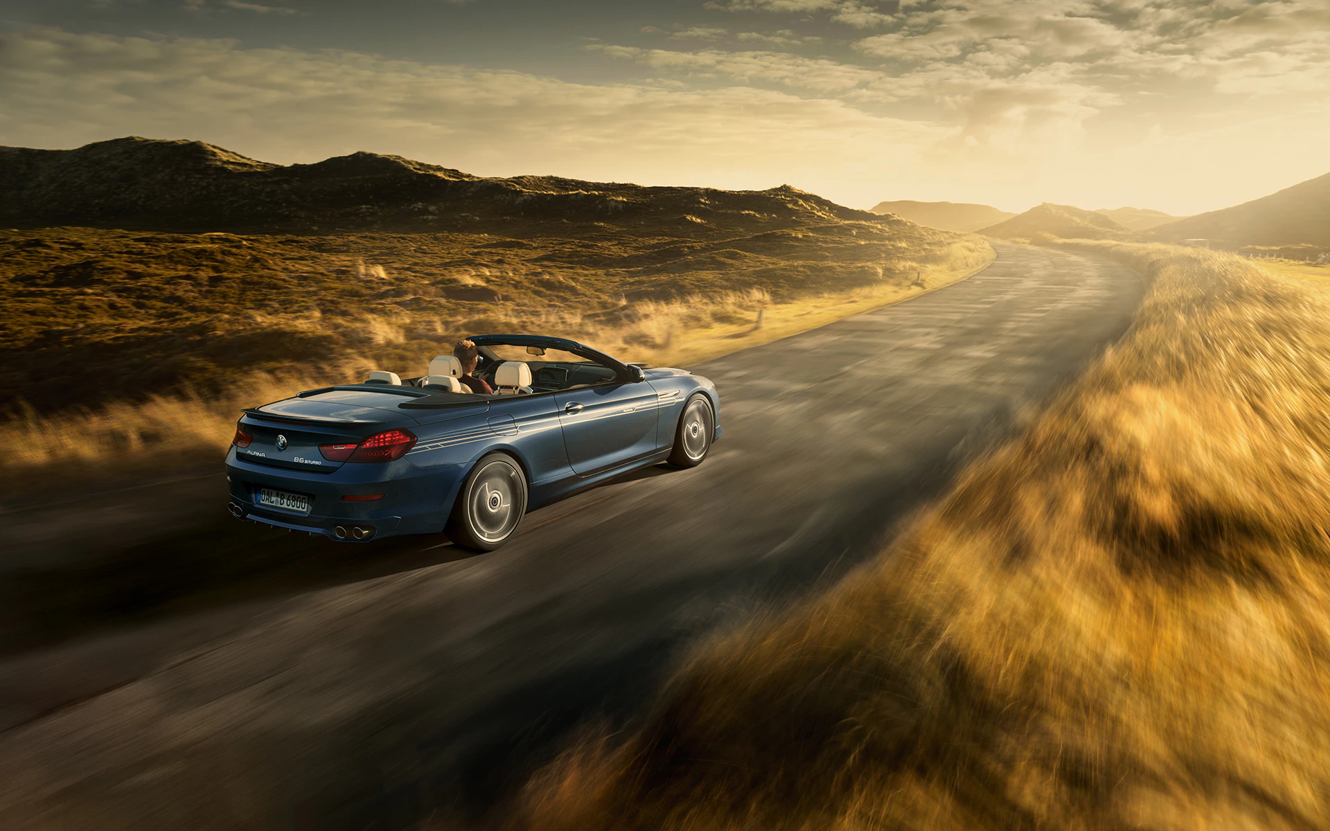 Blaues Cabriolet fährt bei Sonnenuntergang auf einer Landstraße durch eine hügelige Landschaft. Fahrer genießt die Fahrt, umgeben von Natur.