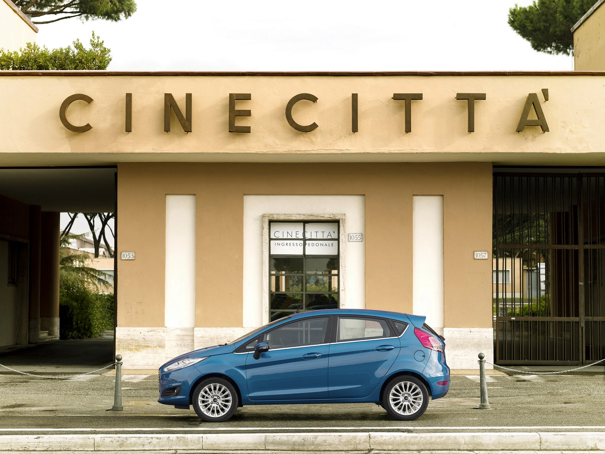 Blaues Auto parkt vor dem Eingang von Cinecittà in Rom, beige Gebäude mit großem Schriftzug, Bäume im Hintergrund.
