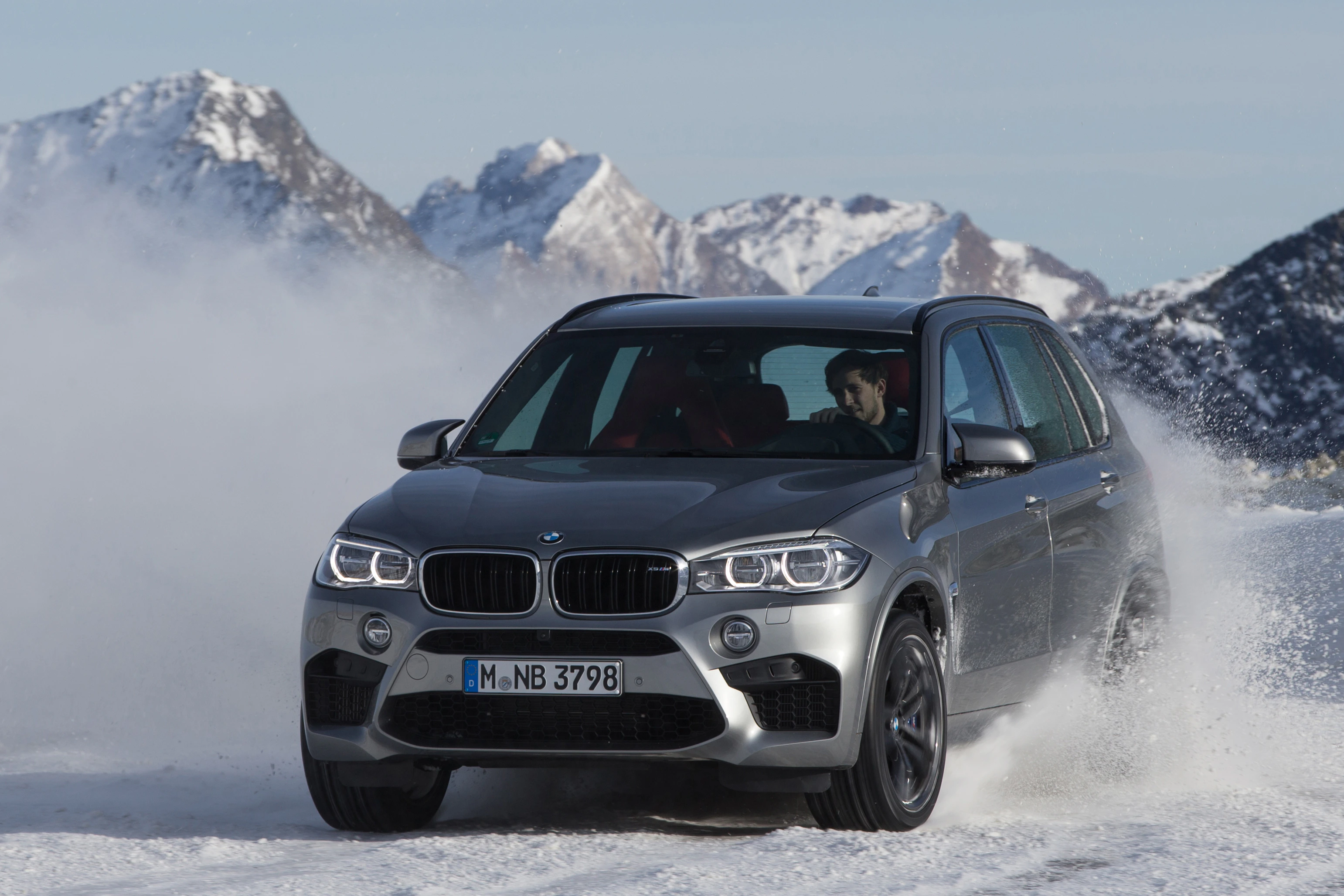 Silberner BMW SUV driftet dynamisch durch Schnee vor verschneiten Berggipfeln. Fahrer im Inneren, klare Winterlandschaft im Hintergrund.