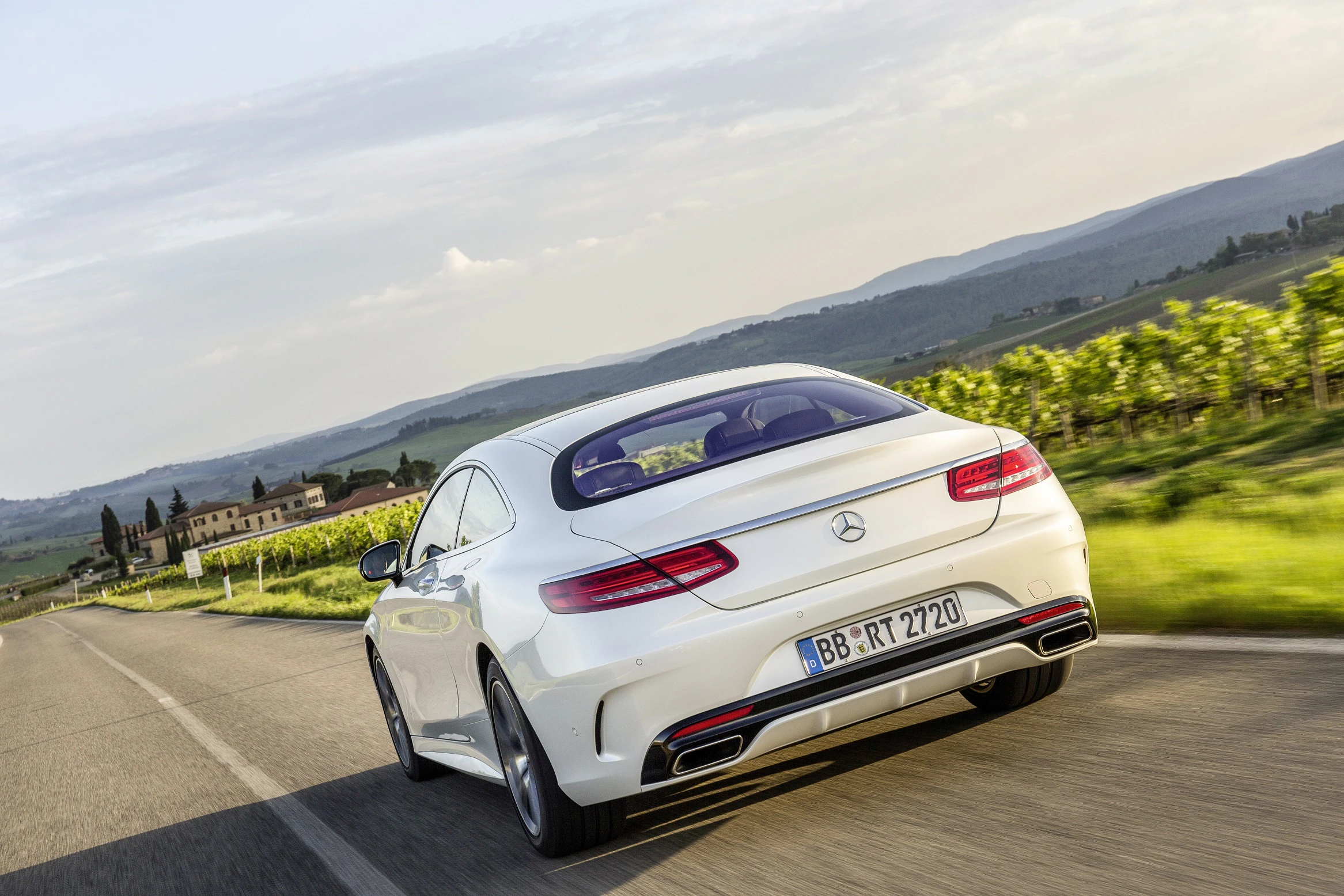 Ein weißer Mercedes-Benz fährt auf einer Landstraße durch eine malerische Landschaft mit grünen Feldern und Hügeln im Hintergrund.