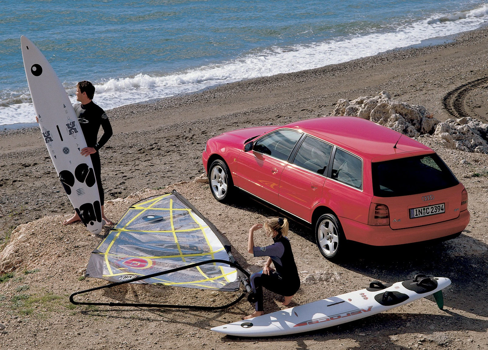 Eine Frau und ein Mann bereiten sich neben einem roten Kombi am Strand auf das Windsurfen vor. Zwei Surfbretter und ein Segel sind sichtbar.