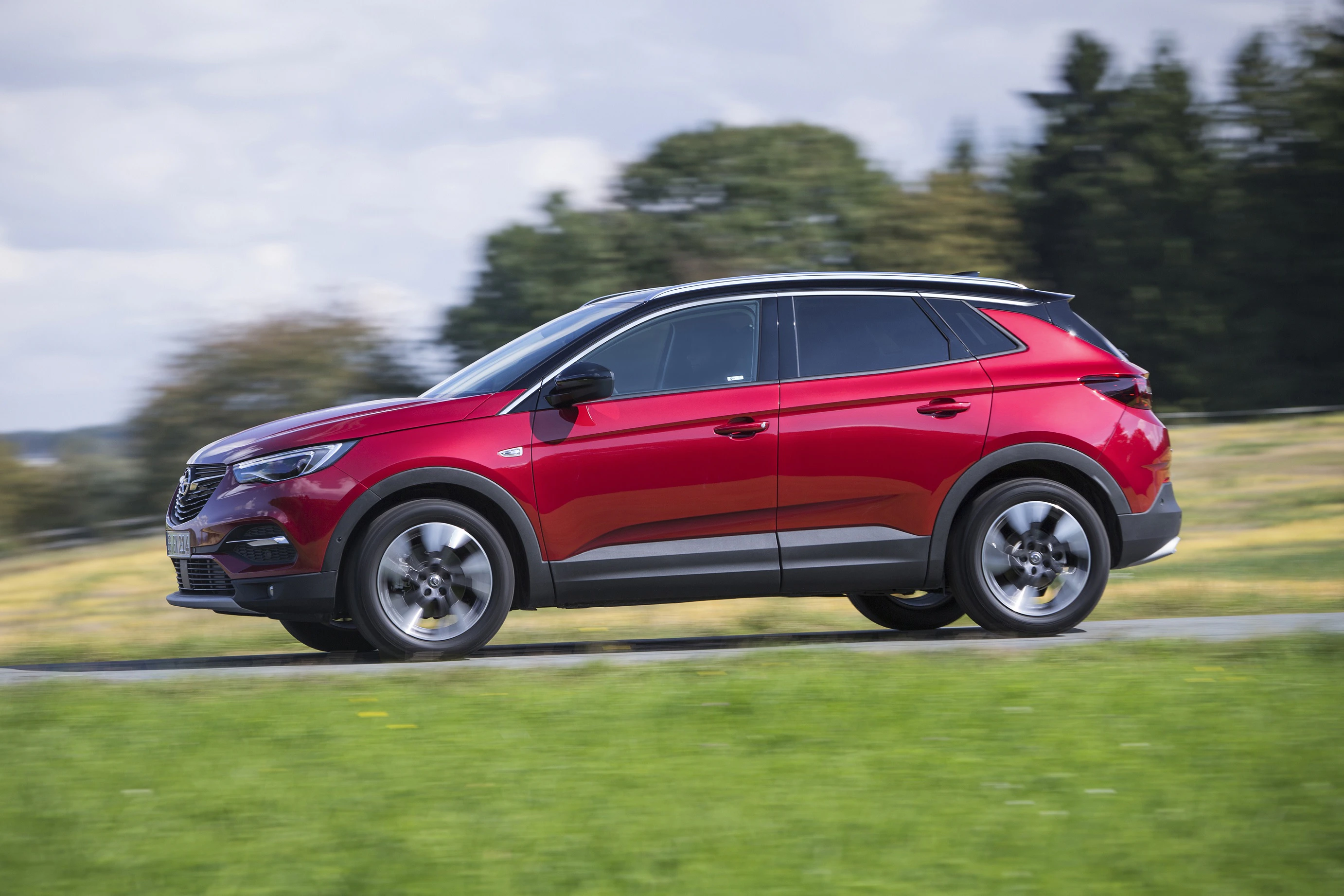 Roter SUV fährt schnell auf einer Landstraße, umgeben von grüner Wiese und Bäumen im Hintergrund unter bewölktem Himmel.