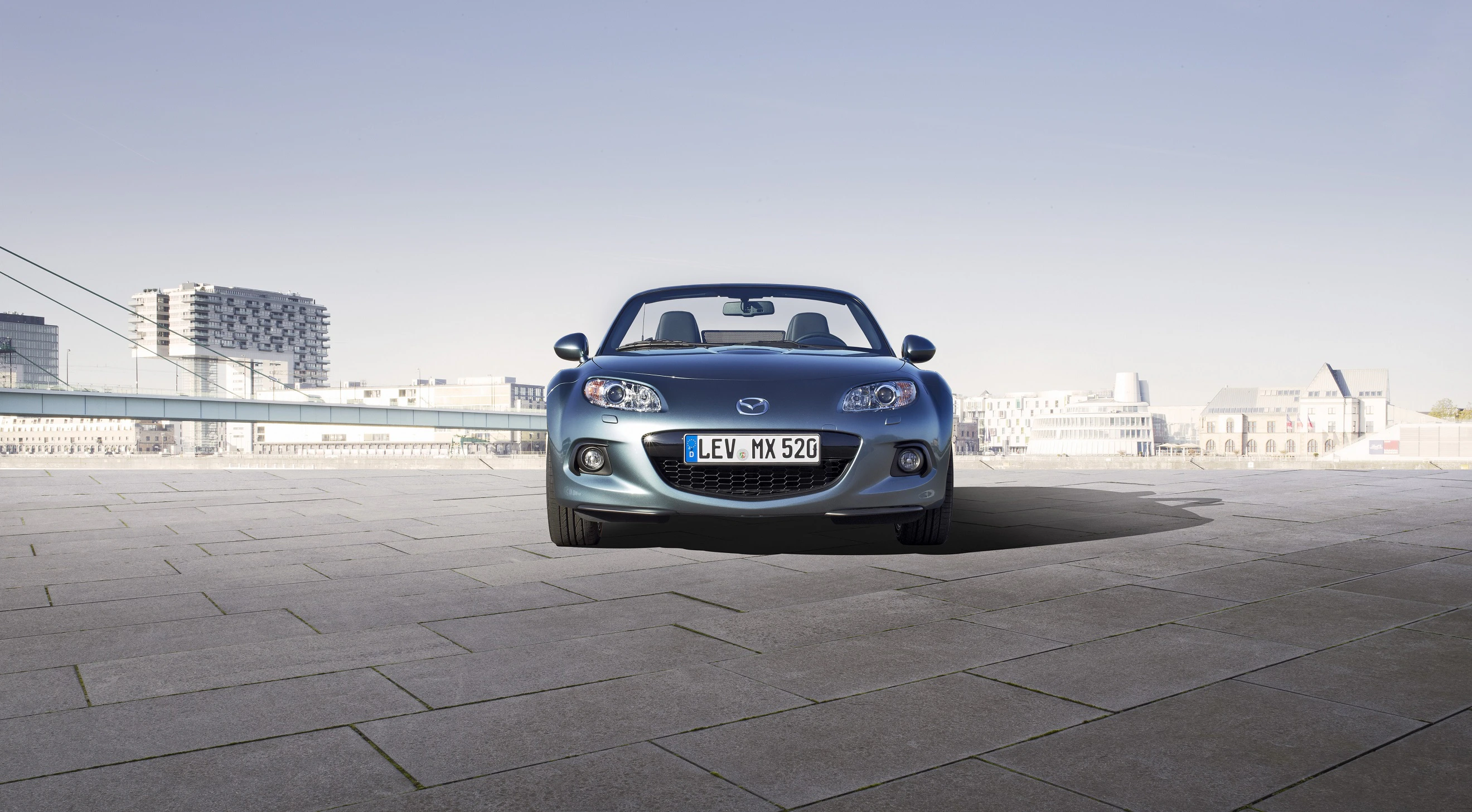 Silberner Cabrio Mazda MX-5 auf einem leeren, gepflasterten Platz, mit modernen Gebäuden im Hintergrund an einem klaren Tag.