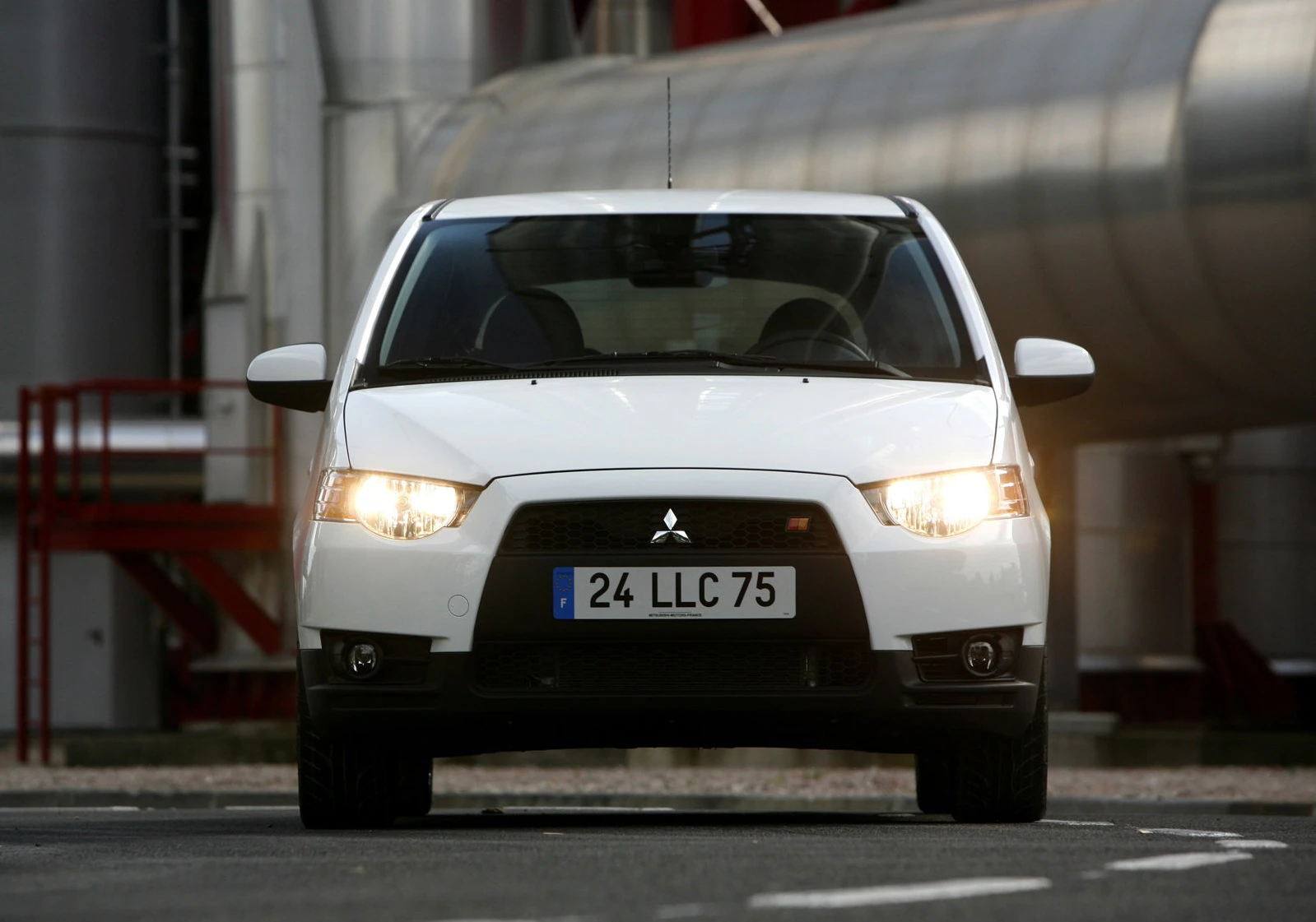 Weißer Mitsubishi Colt Ralliart von vorne, fahrbereit, mit eingeschaltetem Licht und sichtbarem Nummernschild, im industriellen Hintergrund.
