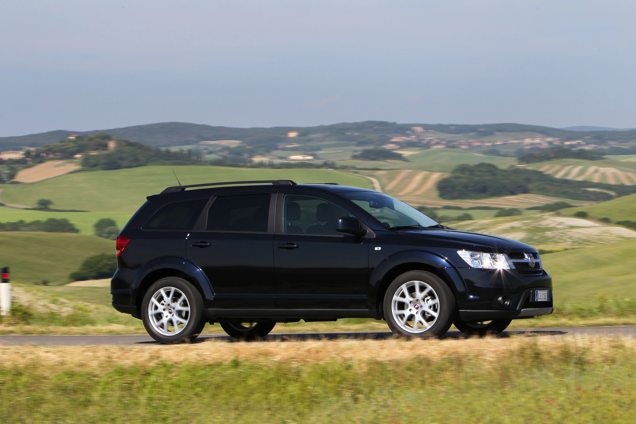 Schwarzer SUV fährt auf einer Landstraße durch hügelige Landschaft, im Hintergrund grüne Felder und blauer Himmel.