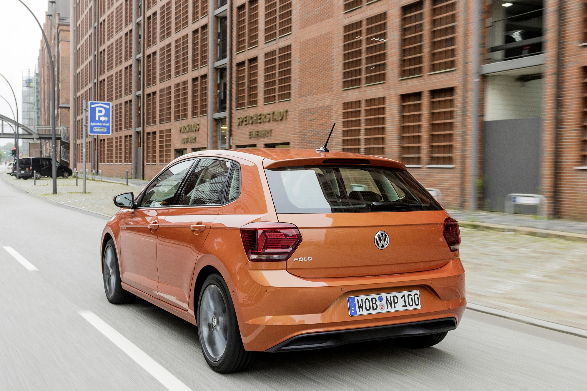 Ein orangefarbener VW Polo fährt auf einer städtischen Straße mit Gebäuden im Hintergrund und einem Verkehrsschild auf der linken Seite.