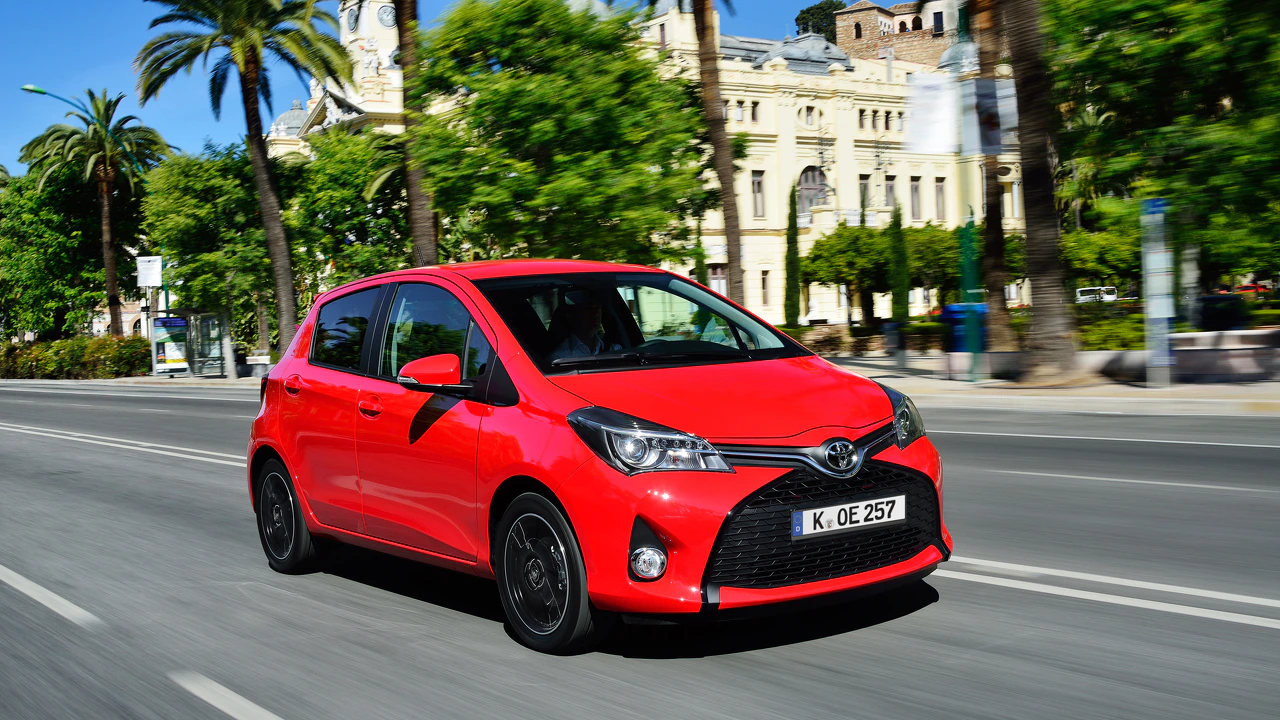 Roter Toyota Kleinwagen fährt auf einer städtischen Straße. Im Hintergrund sind Bäume und Gebäude zu sehen.