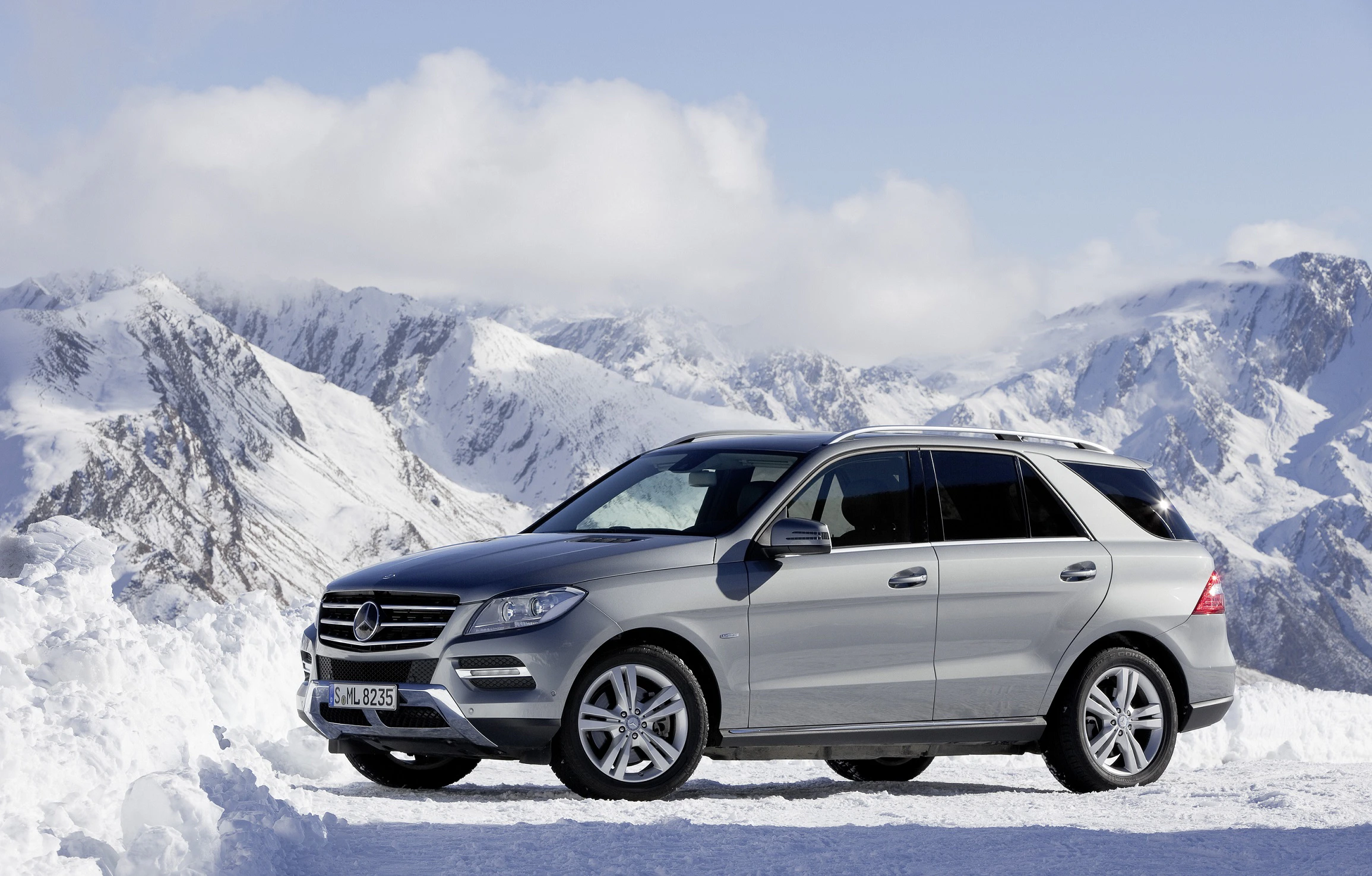 Silberner SUV auf schneebedeckter Landschaft, im Hintergrund majestätische Berge unter bewölktem Himmel.