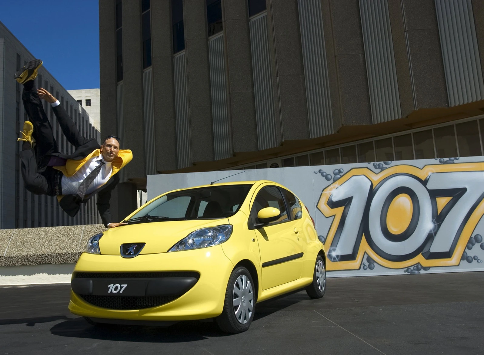 Mann in gelbem Anzug springt dynamisch neben einem gelben Peugeot 107 vor einem Gebäude. Graffiti-Zahl "107" im Hintergrund.