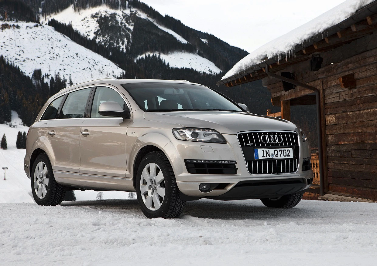 Silberner Audi-SUV parkt im Schnee vor einer Holzhütte, dahinter verschneite Berge und bewaldete Hänge unter klarem Himmel.