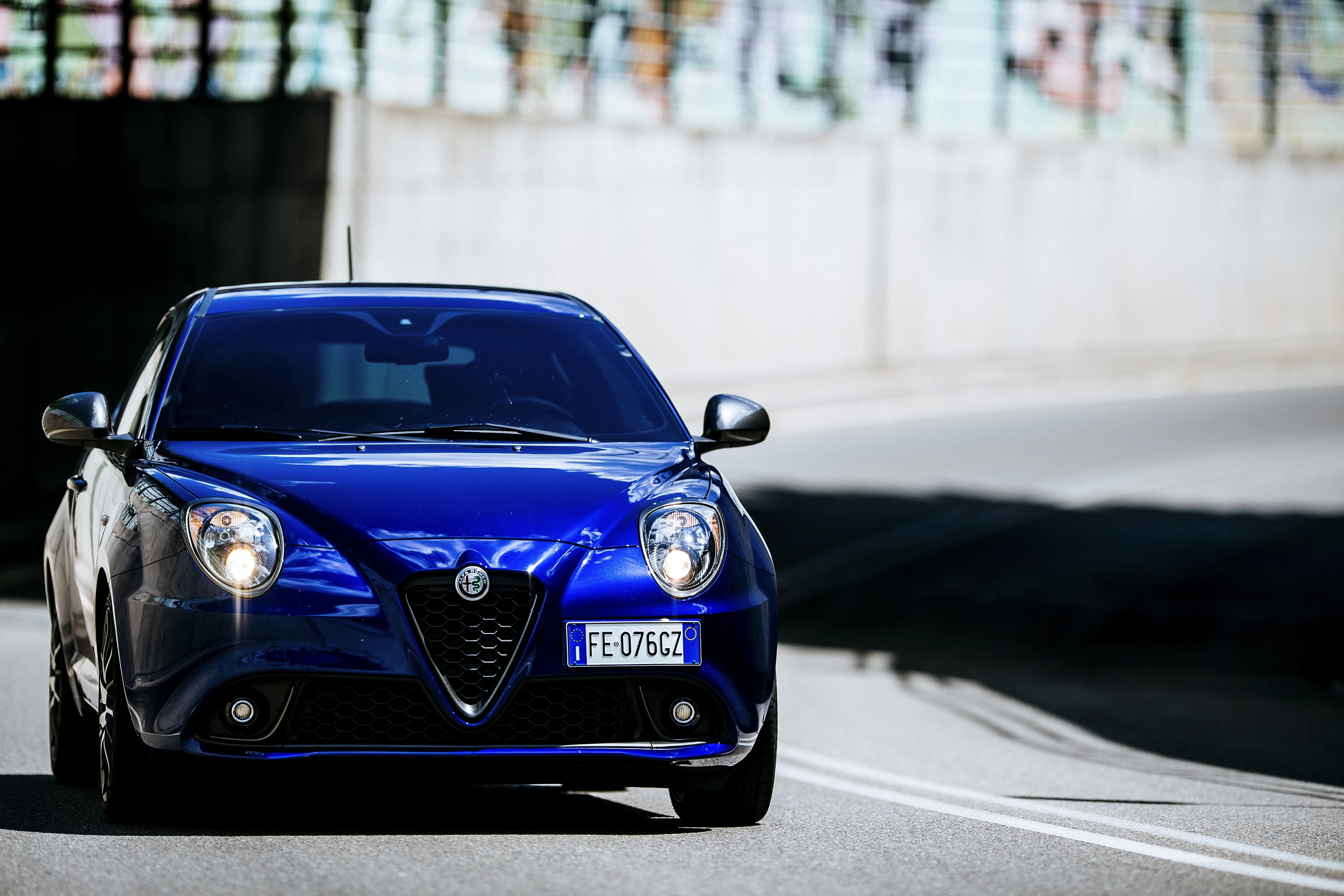 Blauer Alfa Romeo MiTo fährt auf einer städtischen Straße, umgeben von Graffiti an einer Betonwand im Hintergrund.