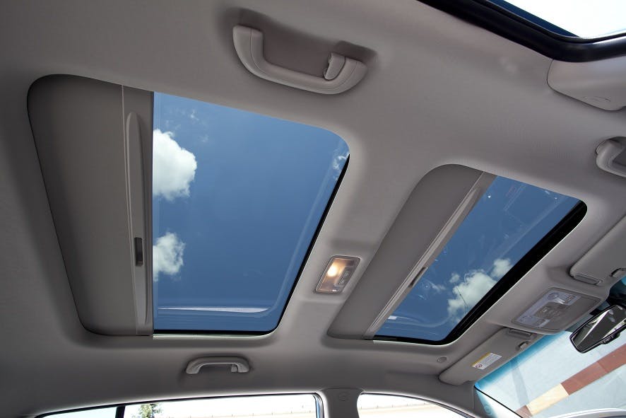 Autodachhimmel mit zwei geöffneten Glasdachfenstern, die blauen Himmel und Wolken zeigen. Innenbeleuchtung und Griffe sichtbar.