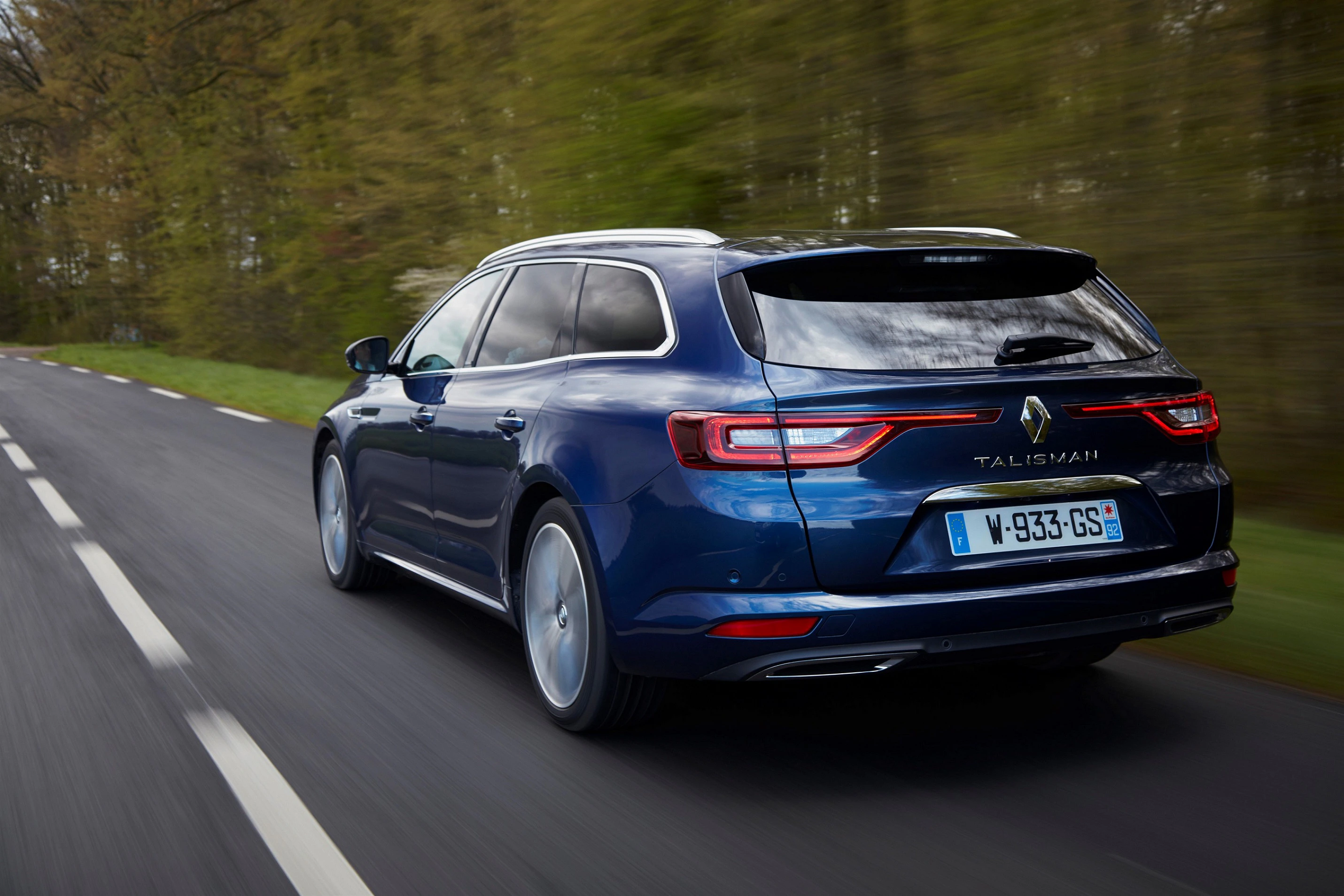 Blauer Renault Talisman Kombi fährt auf einer Landstraße, umgeben von Bäumen. Die Straße ist asphaltiert und leicht verschwommen.